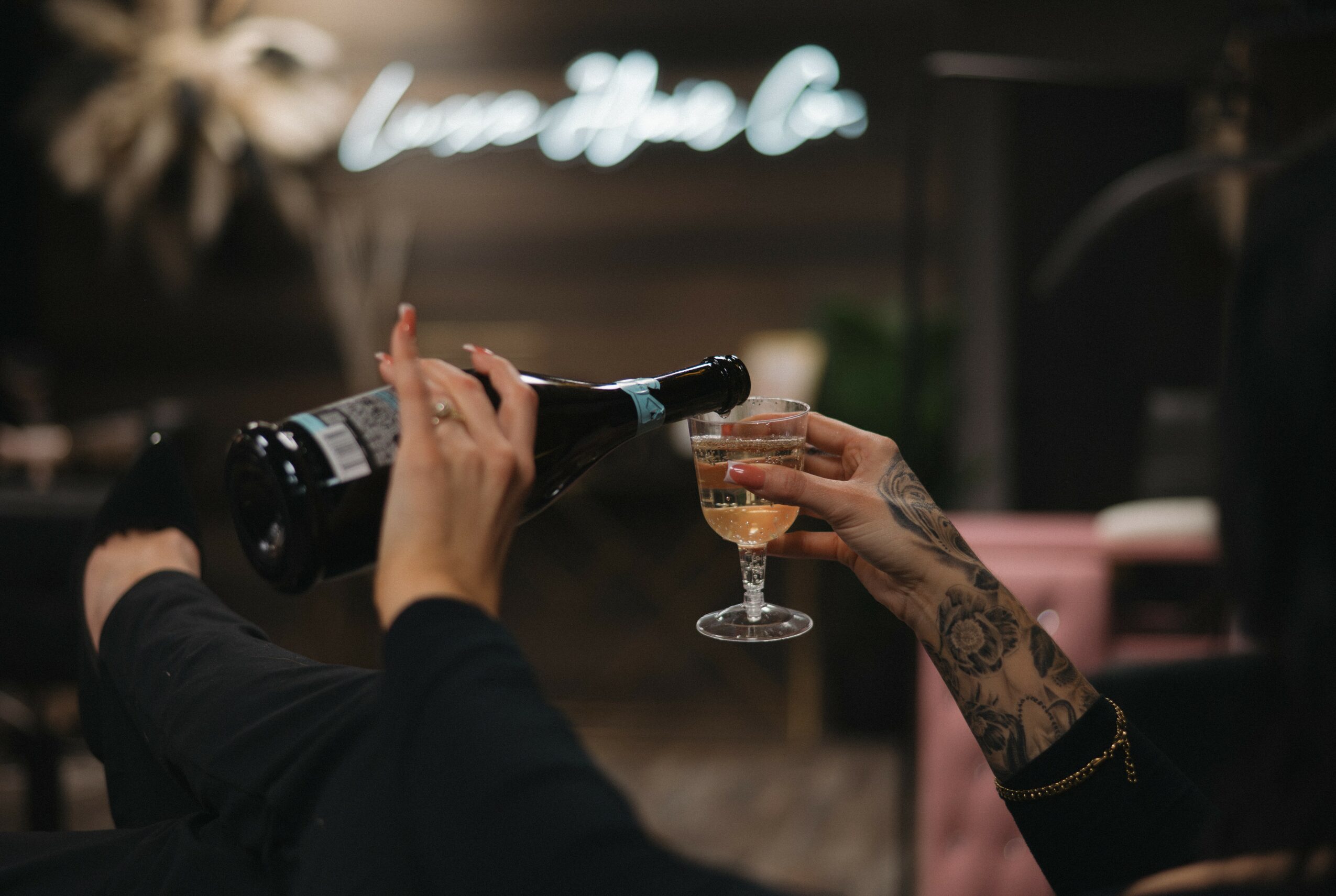 Person pouring wine into a glass held by another person with tattoos, in a dimly lit setting.