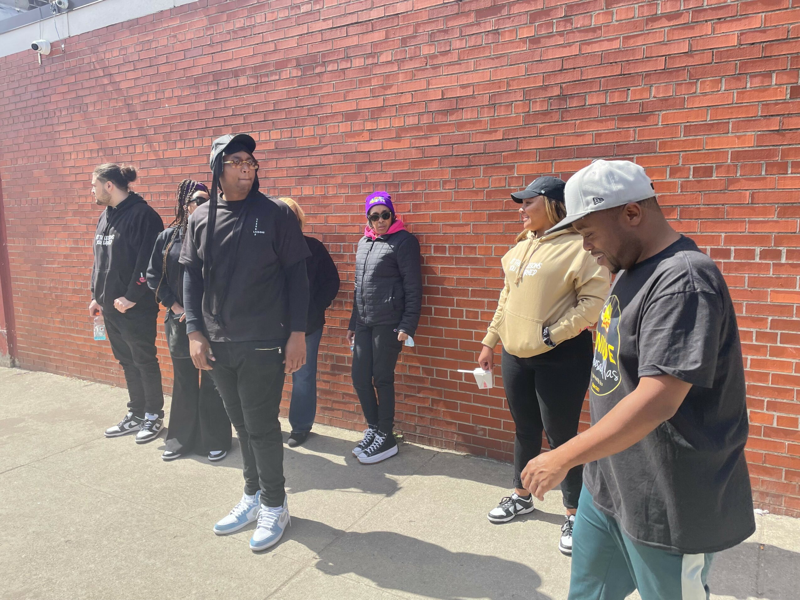 Group of seven people standing in line against a brick wall outdoors, some looking around, some using phones.