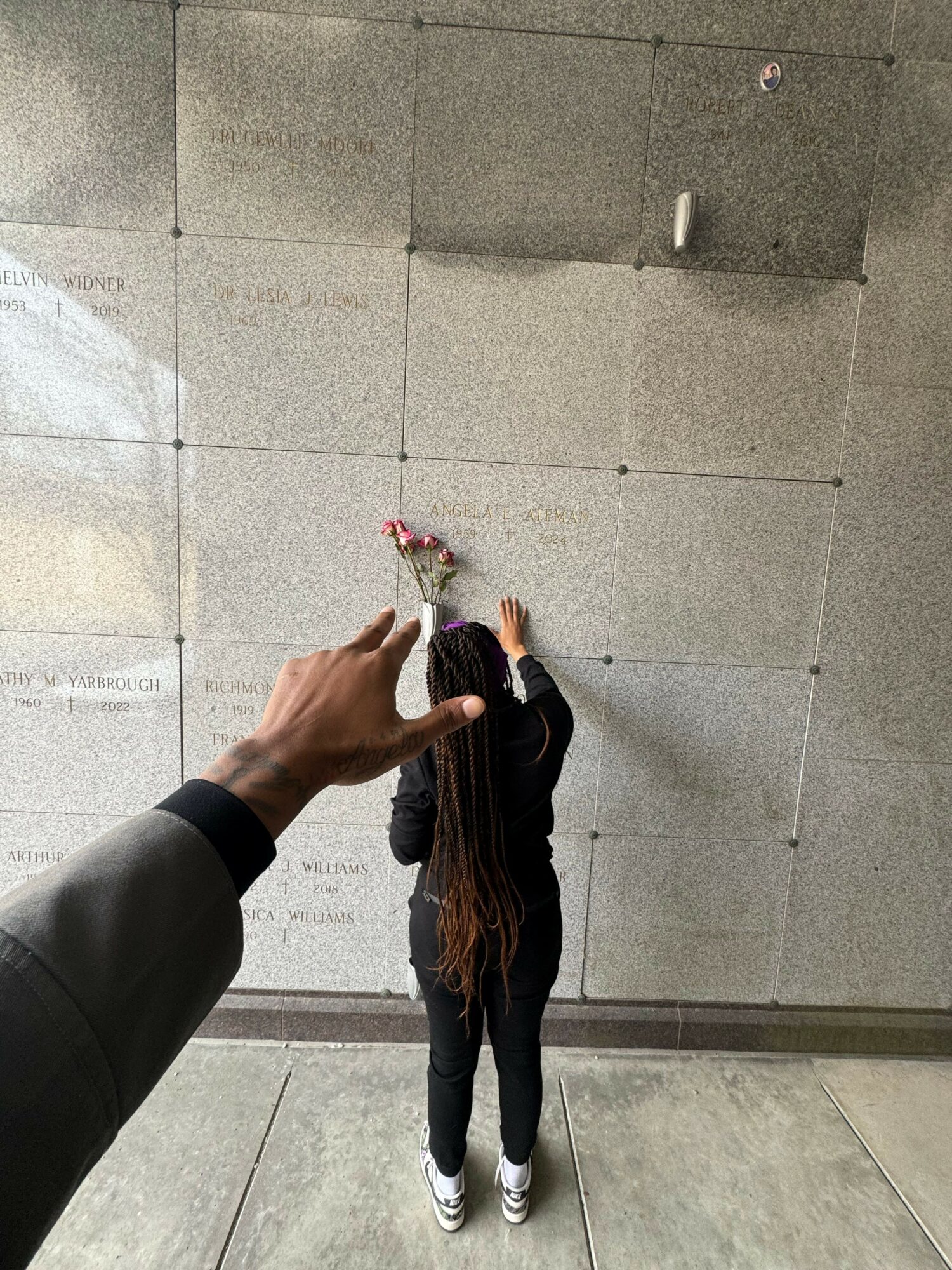 Person holding a small bouquet of flowers, pointing at a woman standing against a tiled wall, with her back to the camera.