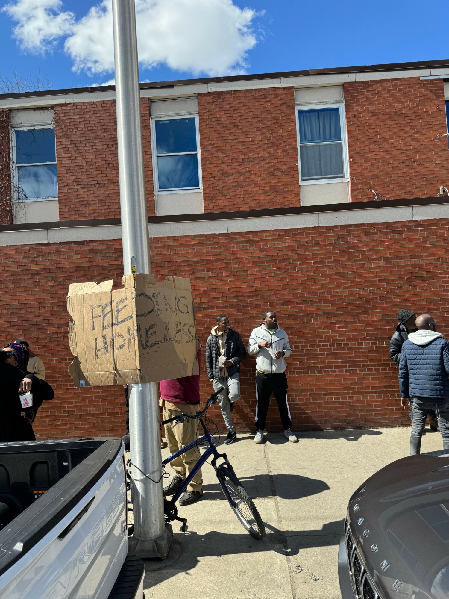 People standing near a bicycle on a sidewalk in front of a brick building, with a cardboard sign attached to a pole.