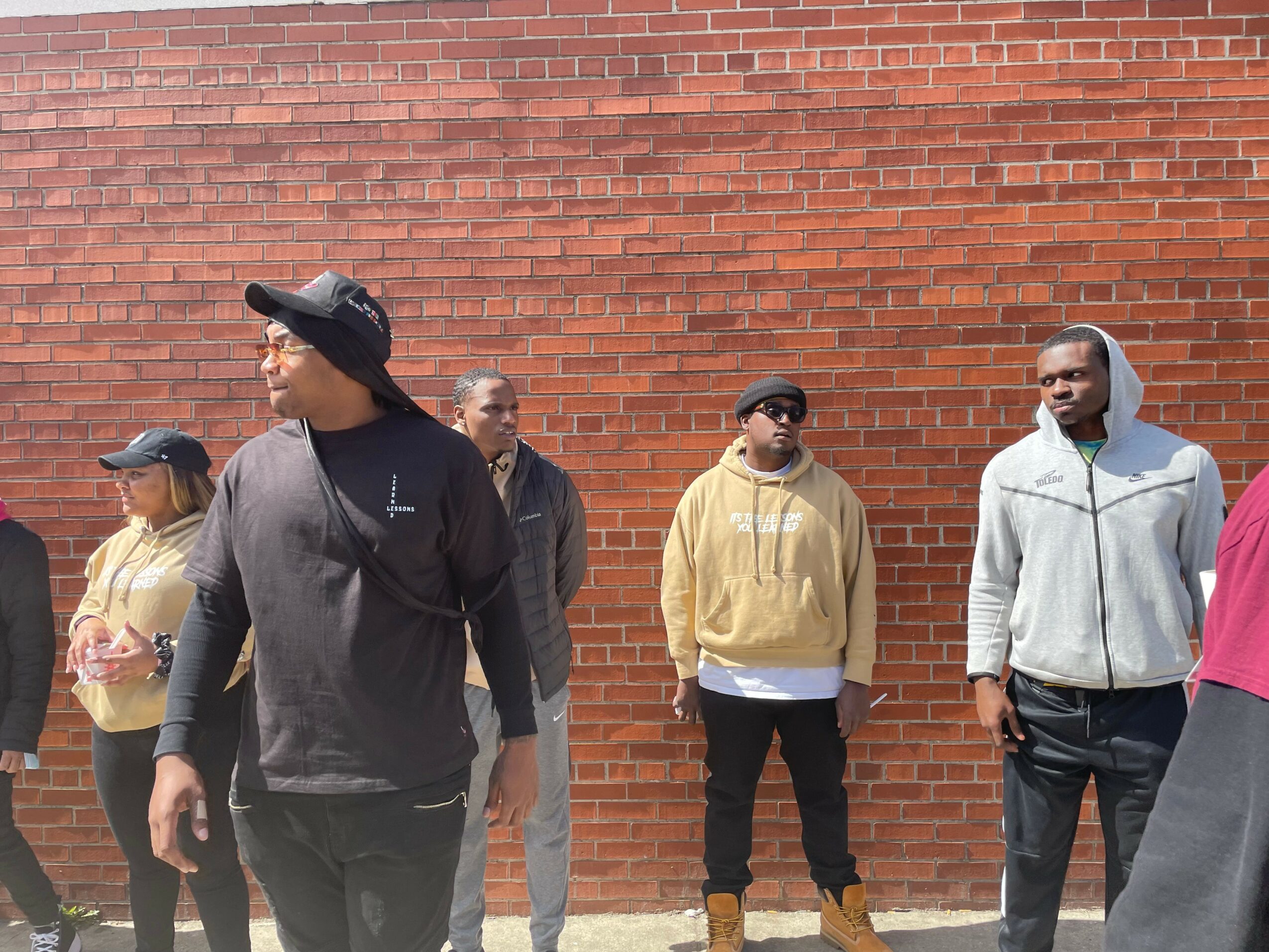 Group of people standing outdoors against a brick wall, some wearing hats and casual clothing.