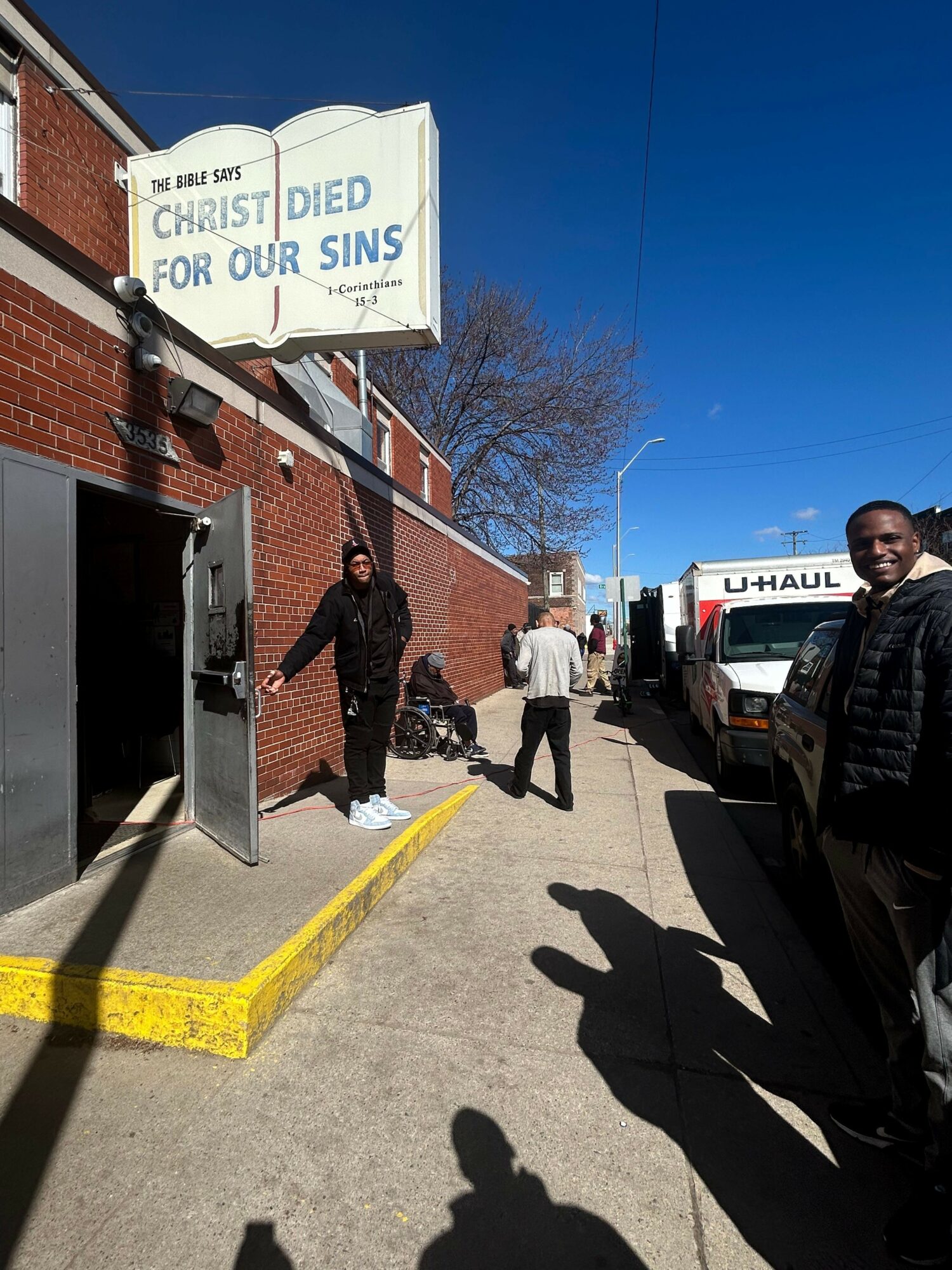 People walking on sidewalk outside a building with a sign, shadows cast on ground, clear blue sky.