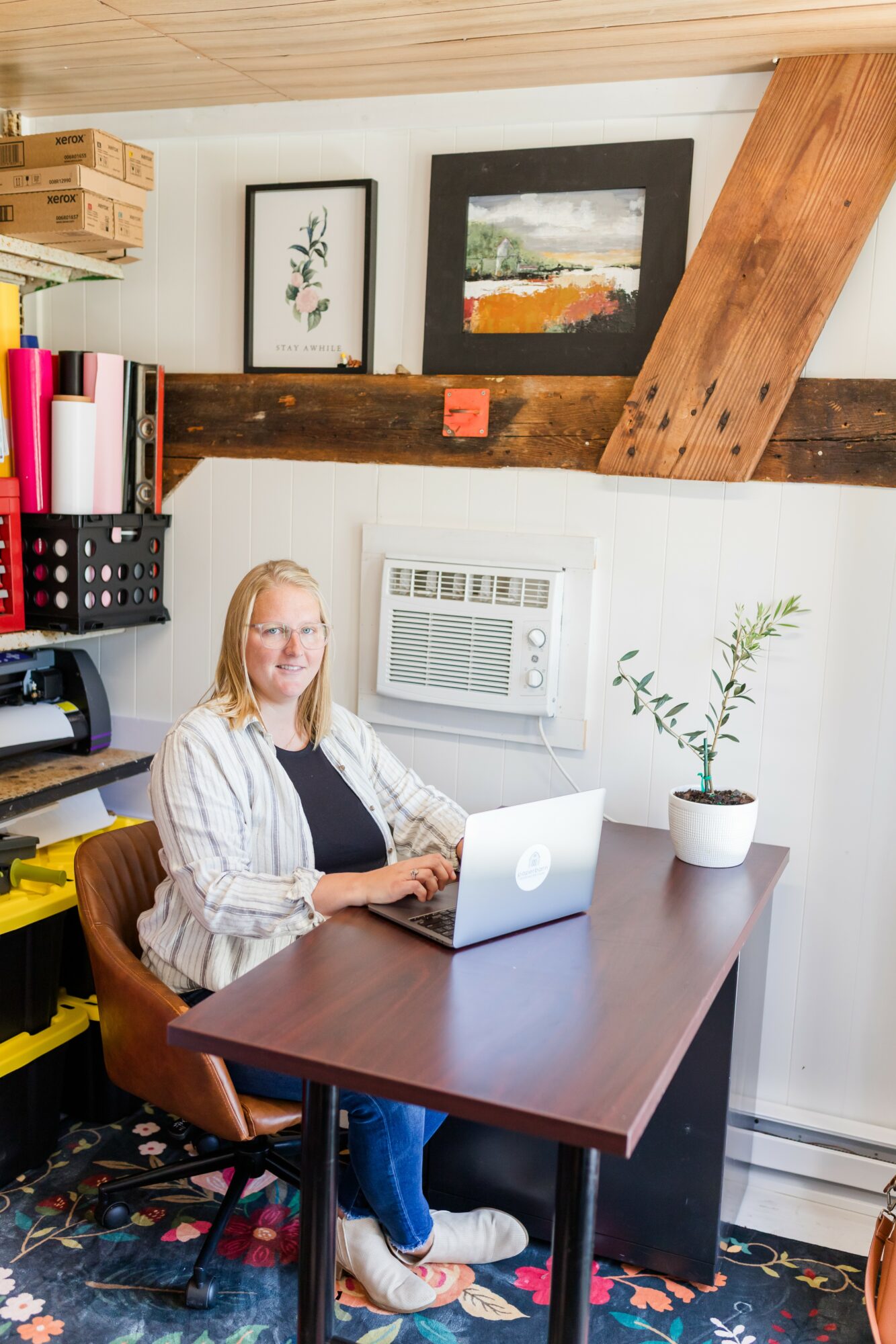 Woman sitting at a desk with a laptop, white wall with framed pictures, potted plant, and colorful storage on wall.