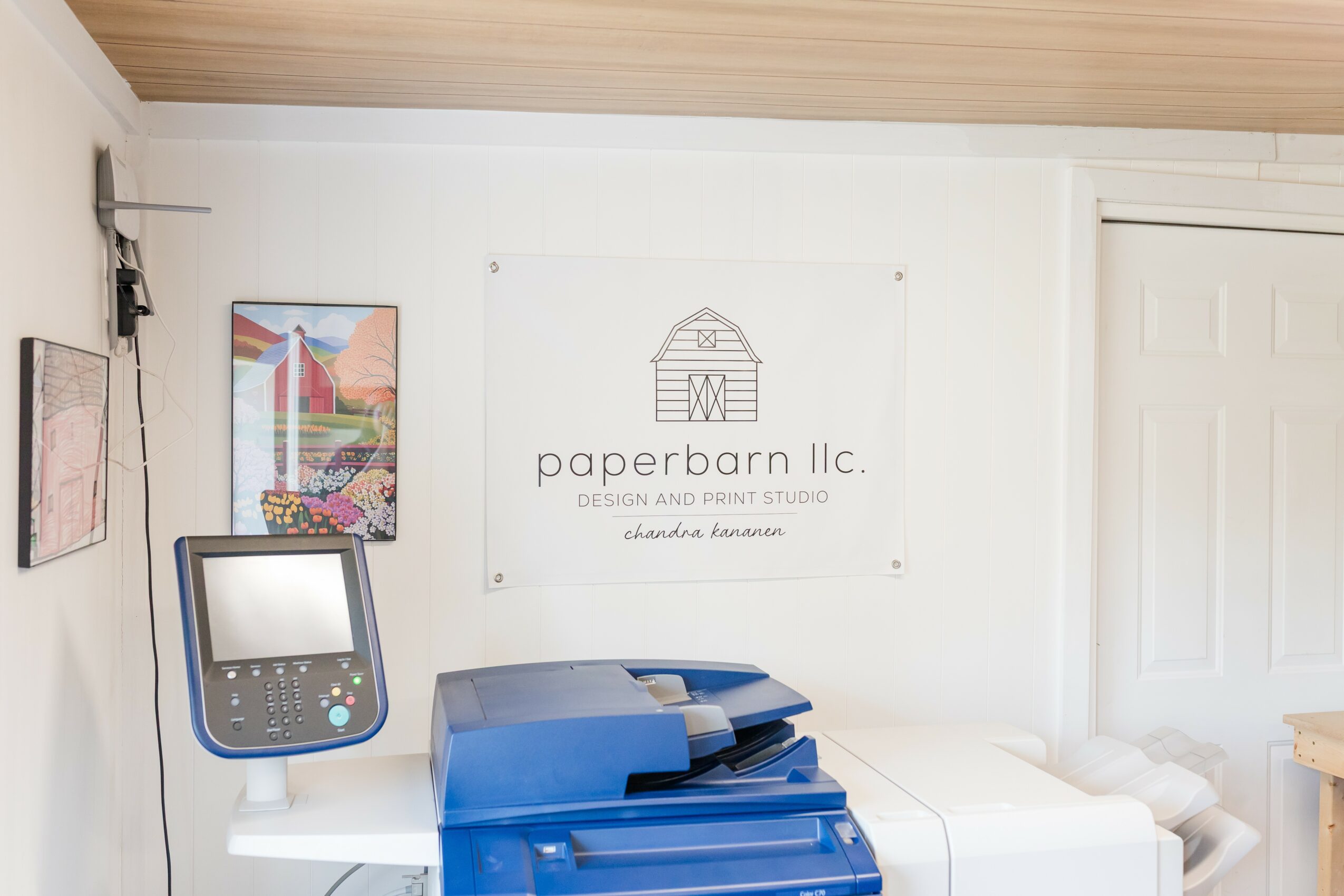 Office space with a white wall, a banner, a printer, and a stack of blue folders, with a door on the right.