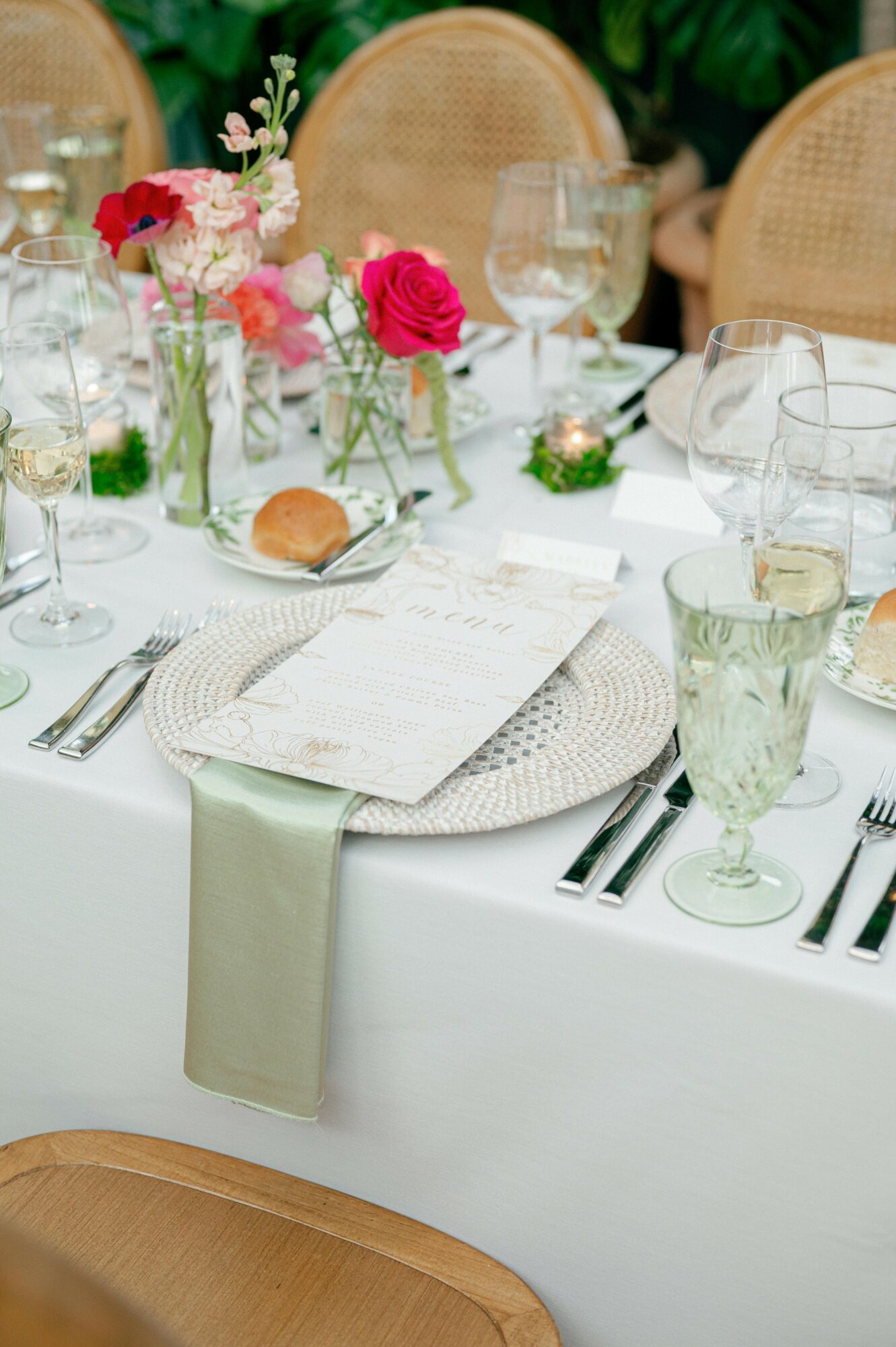 Table setting with flowers, glassware, and a menu, with a white tablecloth and woven placemat, in a dining area.