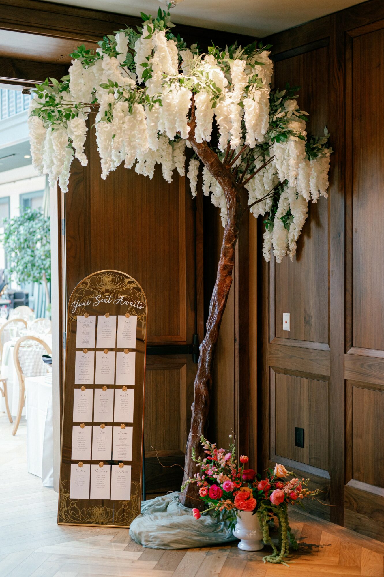 Artificial tree with white cascading flowers and a flower arrangement at base, set indoors against wooden paneling.