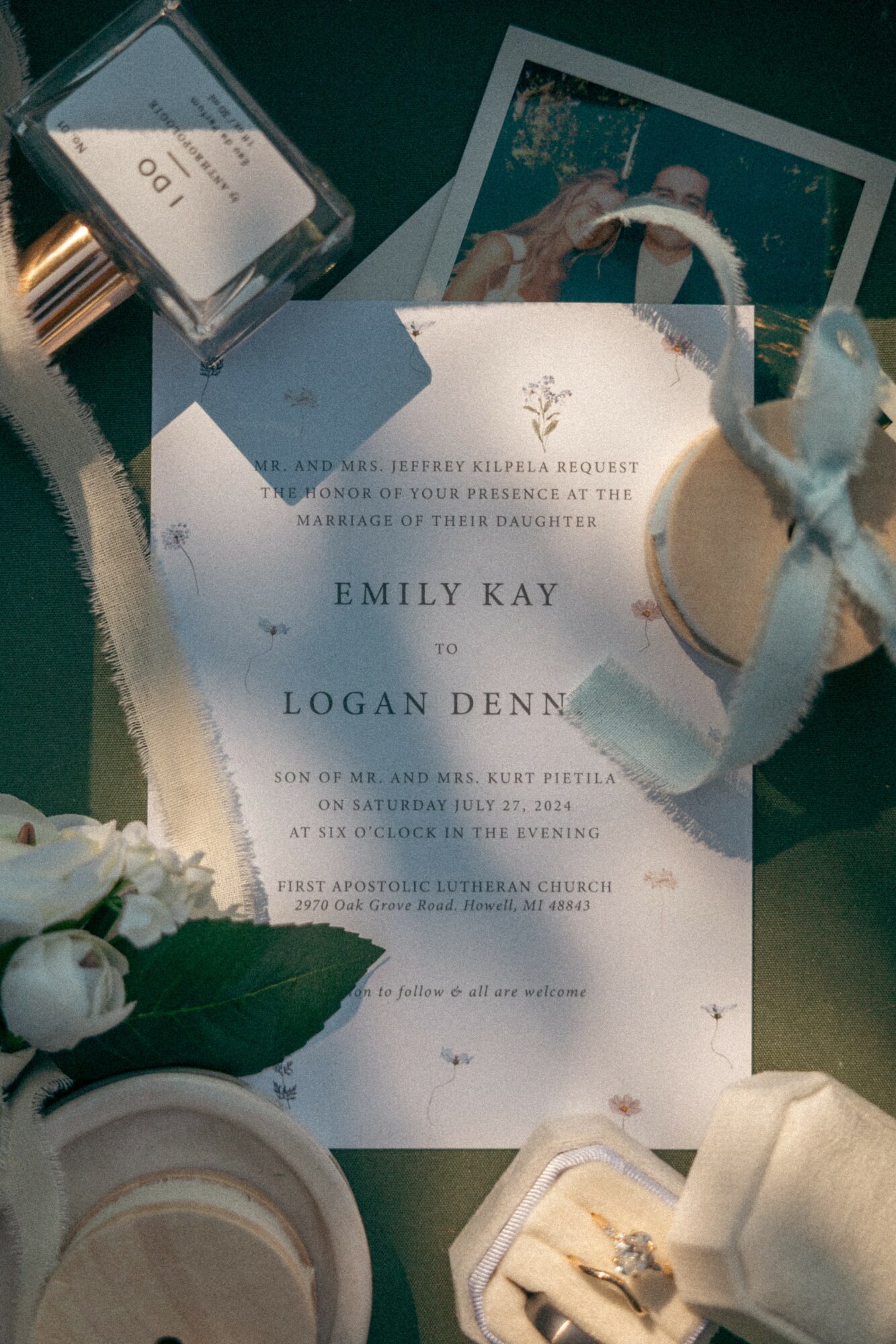Wedding invitation with white flowers, a ring box, and a Polaroid photo on a dark surface.