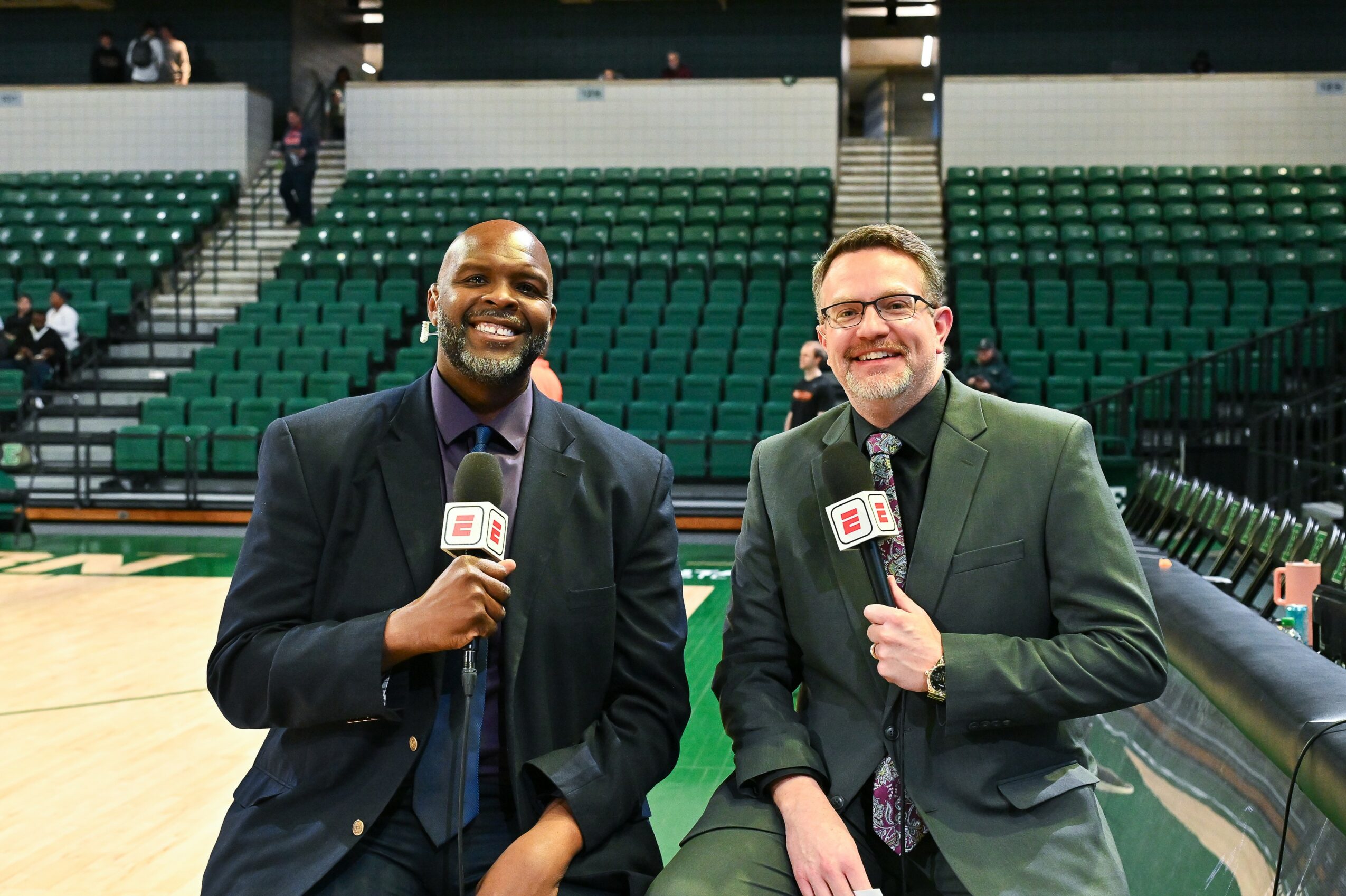 Two men in suits holding microphones stand on a basketball court with empty green seats behind them.