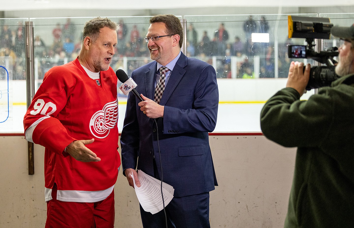 Person in red hockey jersey being interviewed by a man in a suit, with a cameraman recording the scene, on an ice rink.