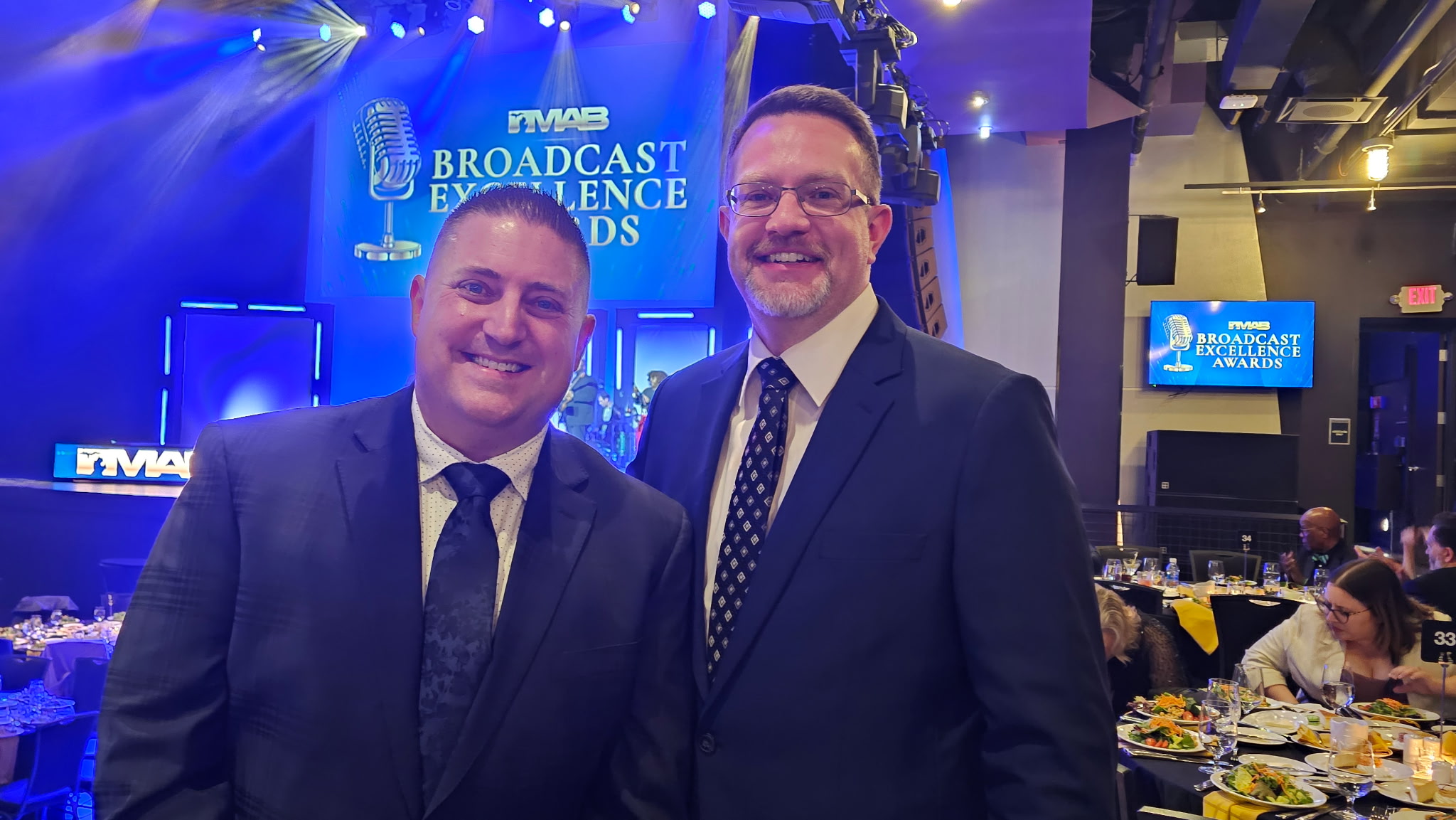 Two men in suits smiling at an event with a stage and screens in the background.