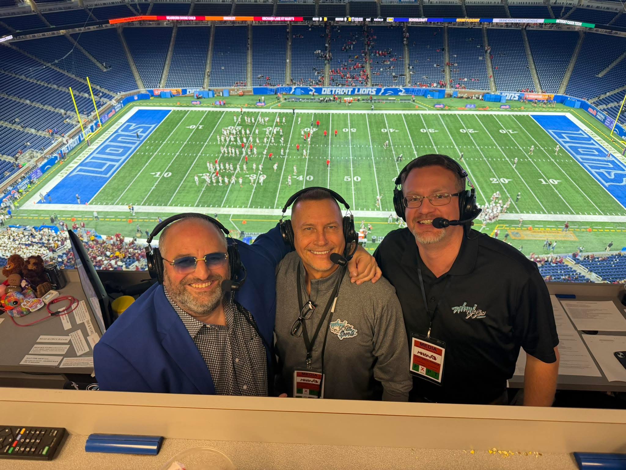 Three men wearing headsets and badges sit in a stadium press box with a football field in the background.