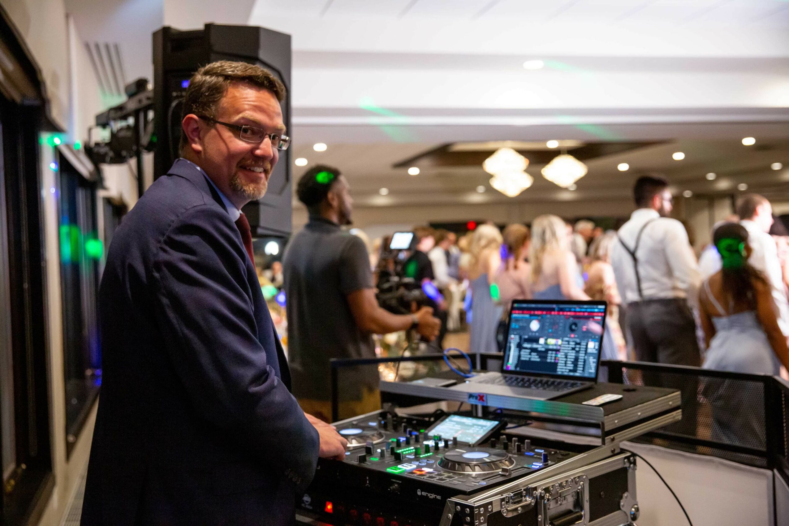 Man with glasses and beard smiling at DJ equipment in a crowded event space, people in background, laptop open, colorful lighting.