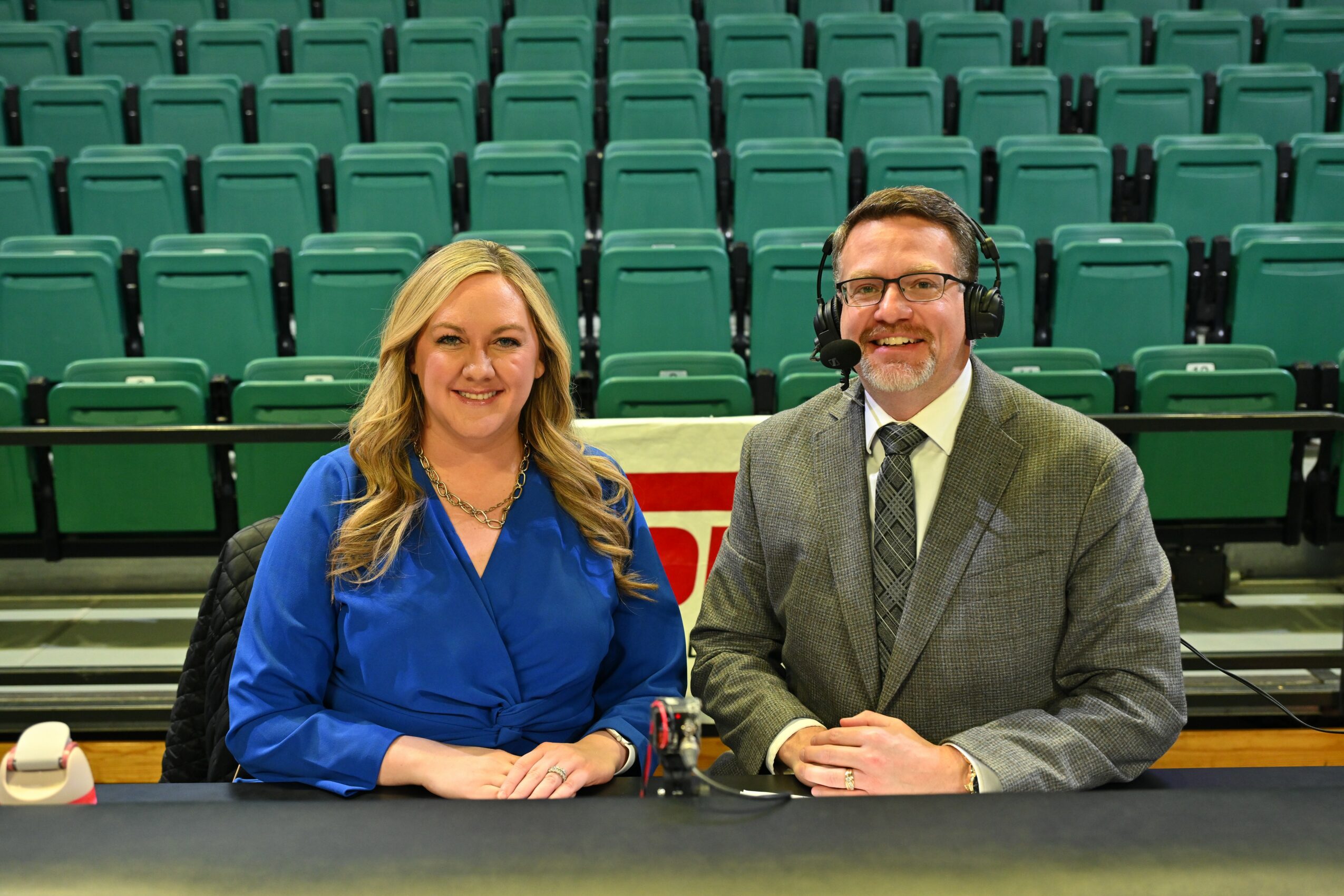 Two people sit at a table in front of empty green seats, smiling, with a microphone between them.