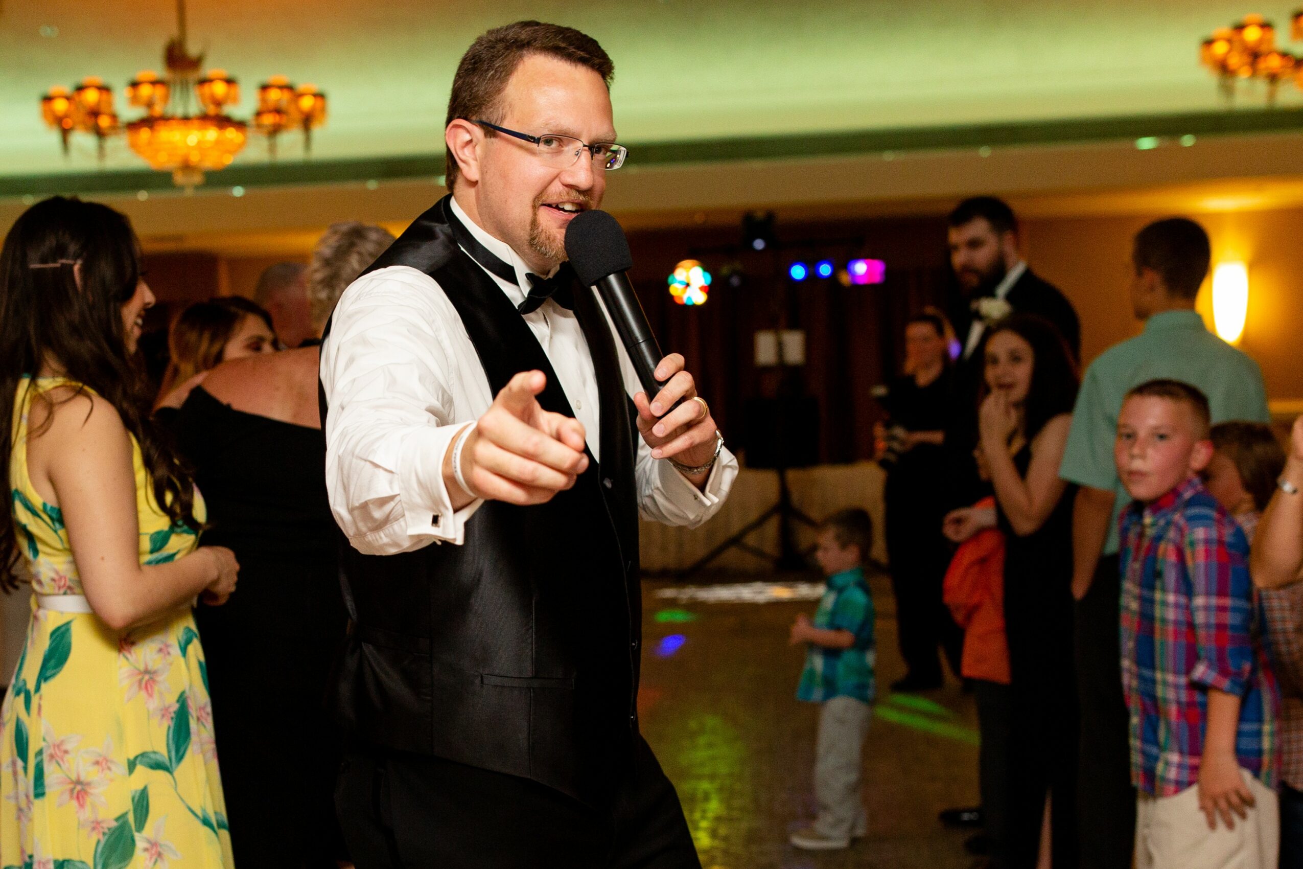 Man in tuxedo holding microphone, pointing, at a party with children and adults in background.