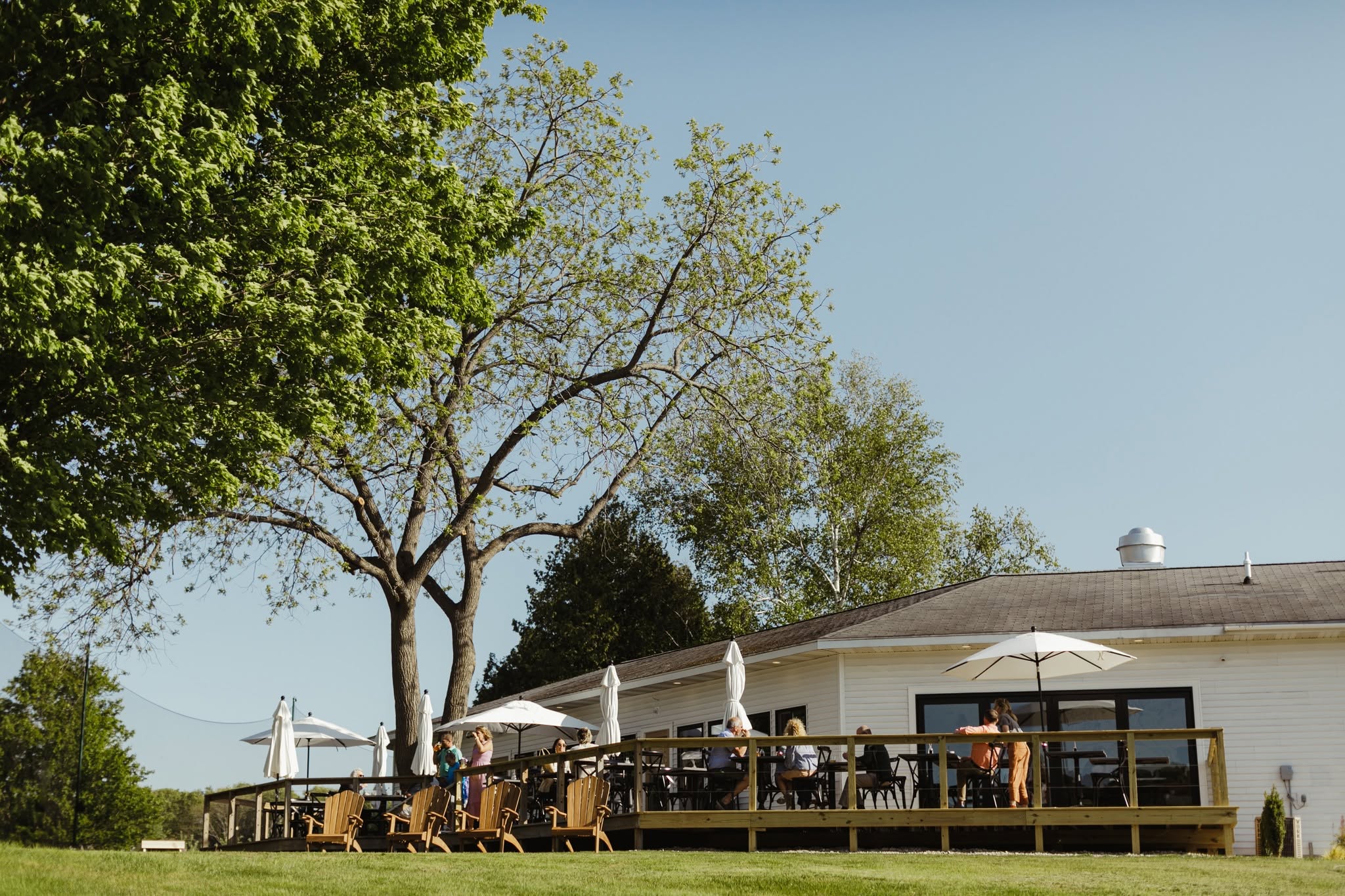 Outdoor patio with tables, chairs, and umbrellas near a building, trees in the background, clear sky.
