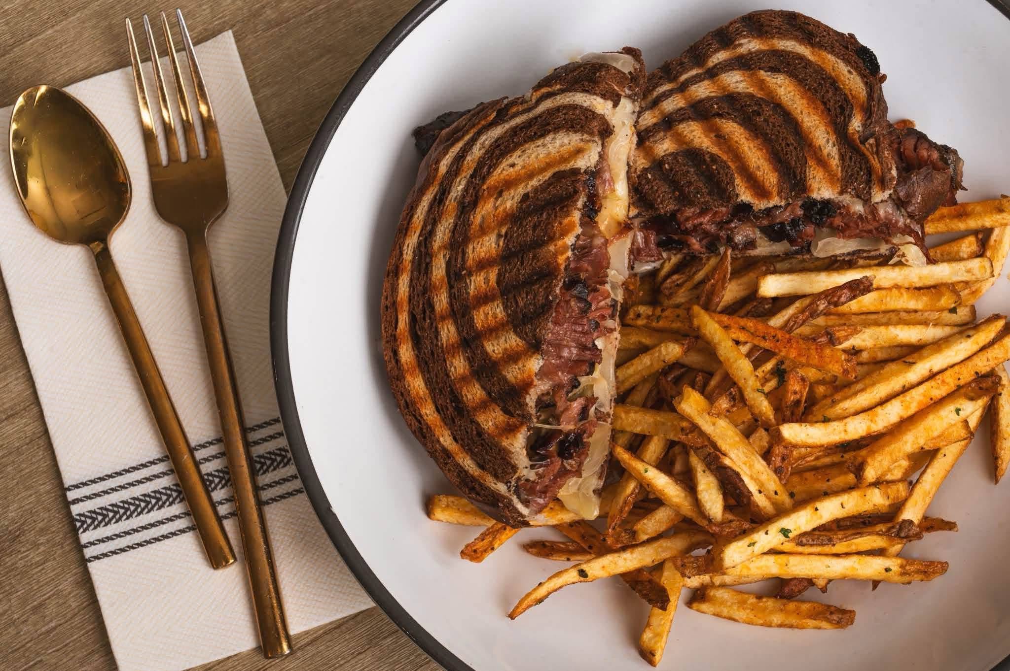 Grilled steak with French fries on a white plate, gold fork and spoon beside a napkin, on a wooden table.