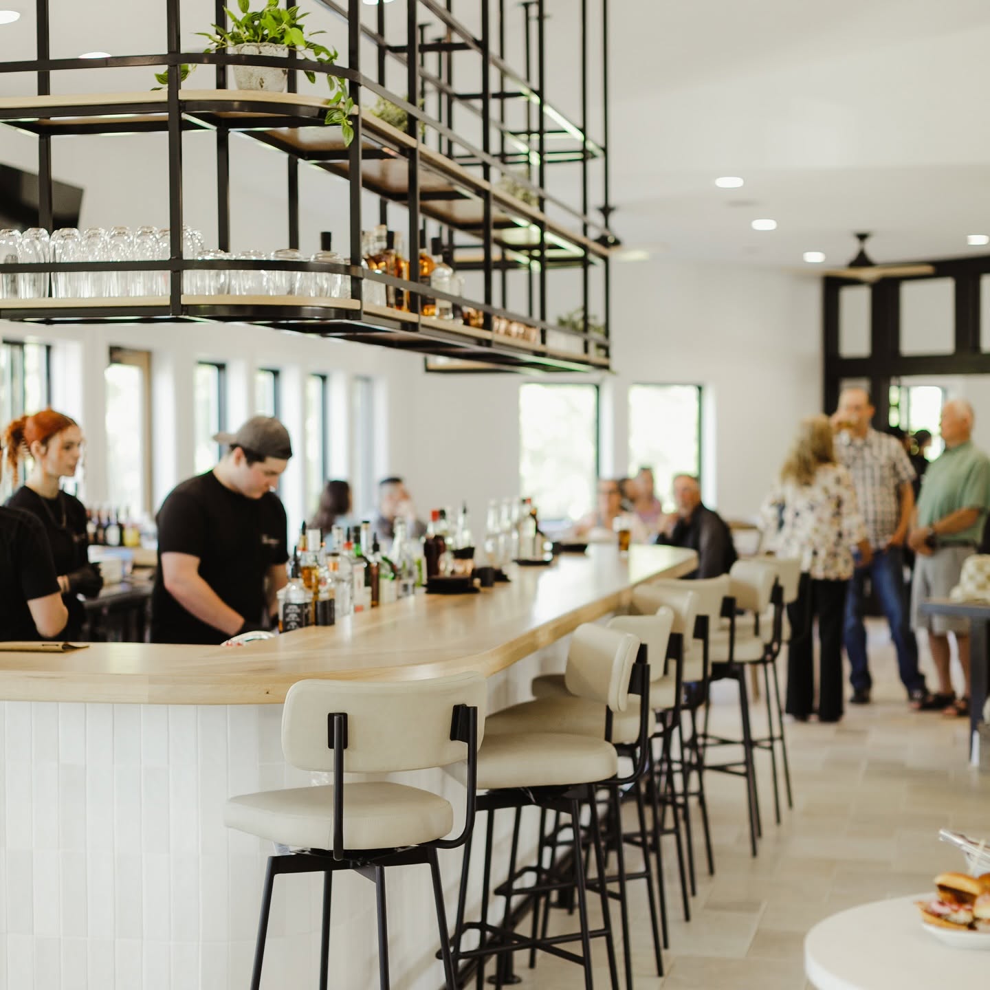 Bar with stools, people socializing, and a modern interior with shelves and large windows.
