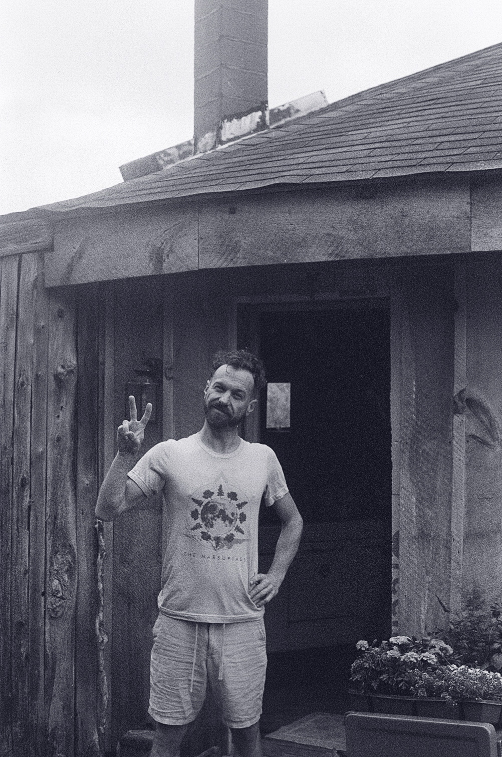 Man with a beard making a peace sign, standing outside a wooden building, wearing a t-shirt and shorts.