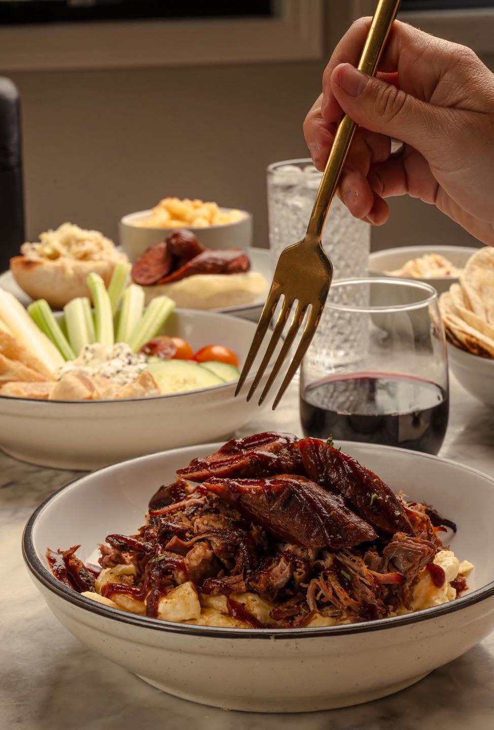 Hand holding a fork over a bowl of shredded meat, with various dishes and a glass of red wine in the background.