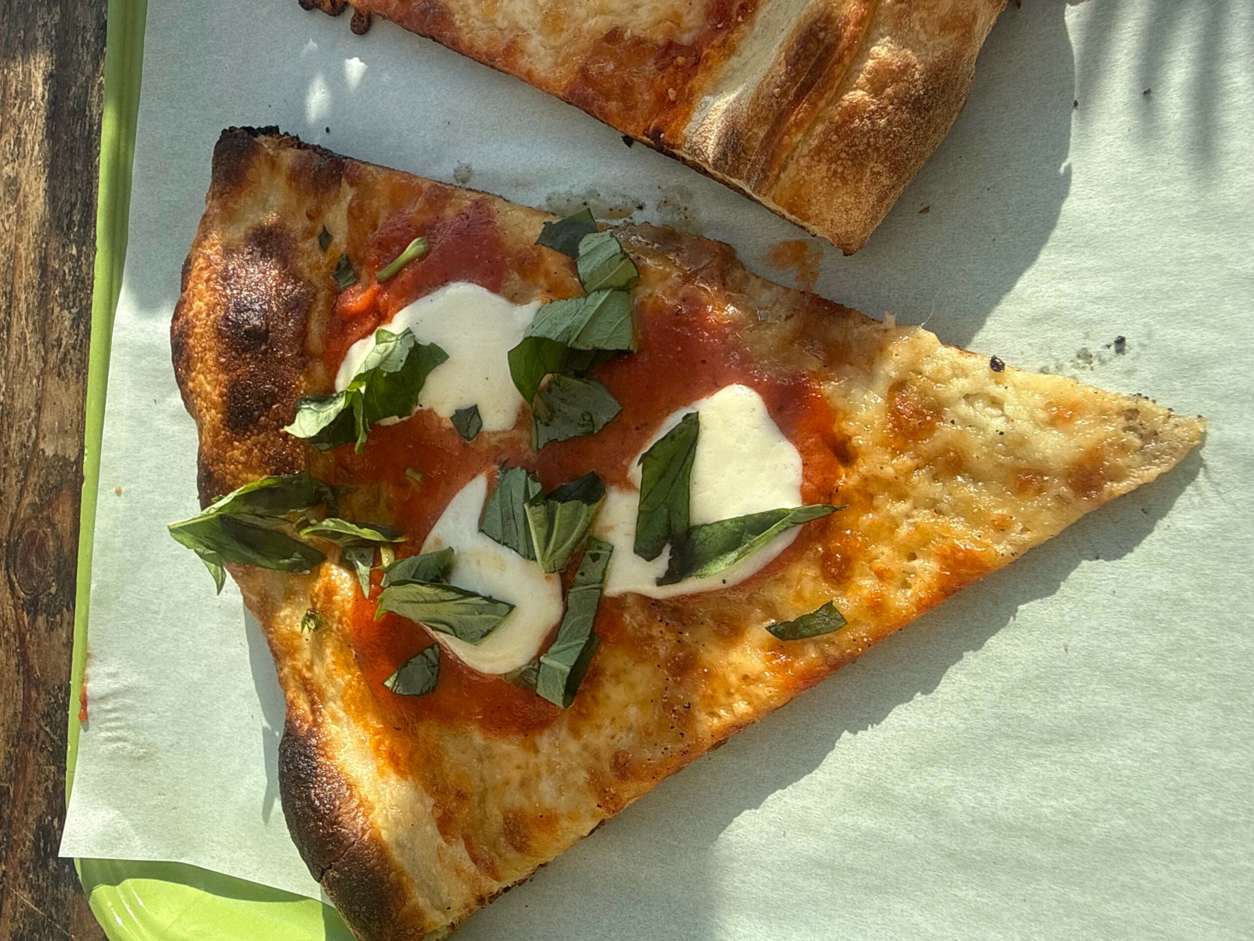 A slice of pizza topped with cheese, tomato sauce, fresh basil, and slices of garlic on a light surface.