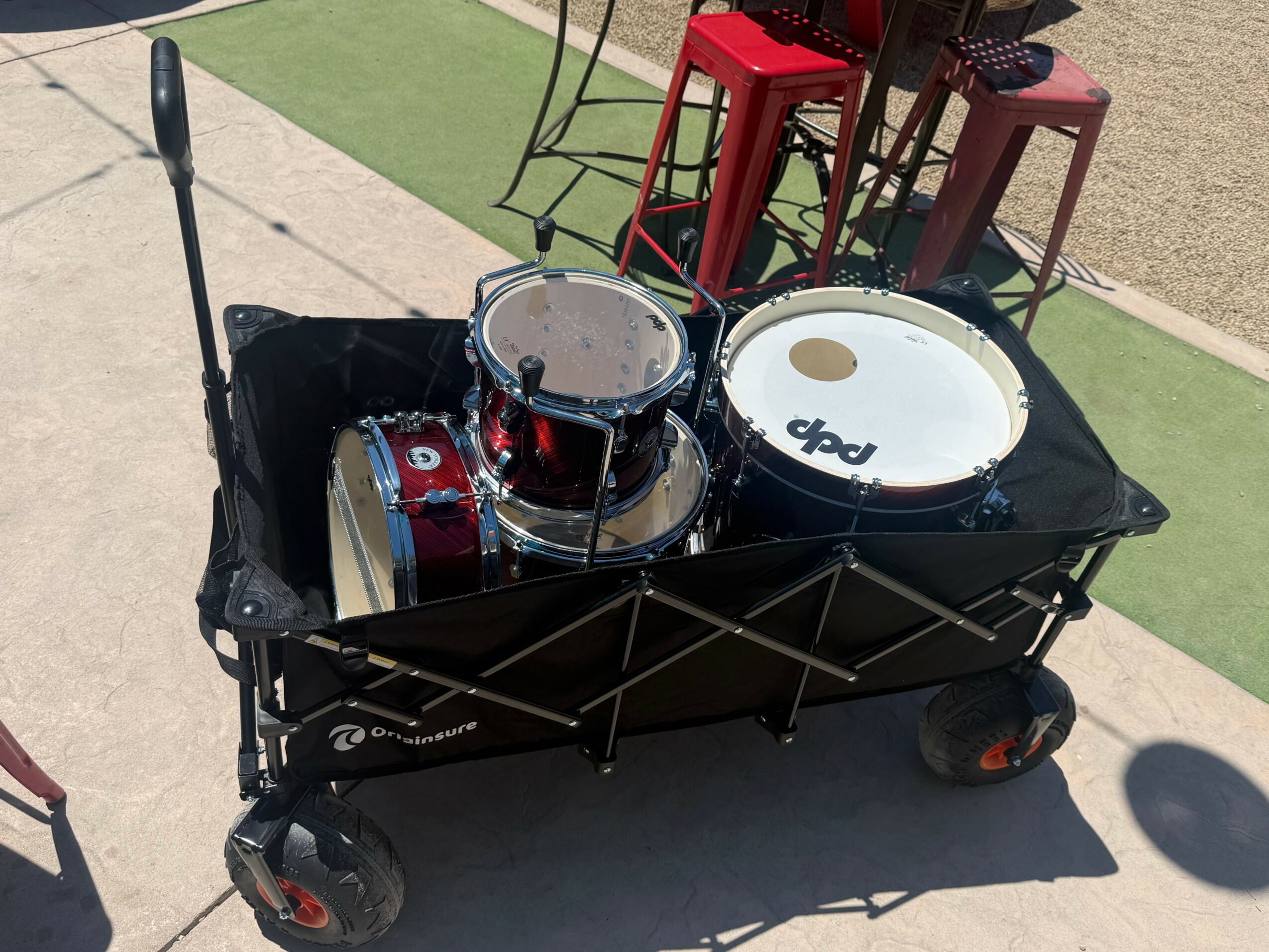 A black wagon with a red drum set inside, including a snare drum and a bass drum, on a sidewalk outdoors.