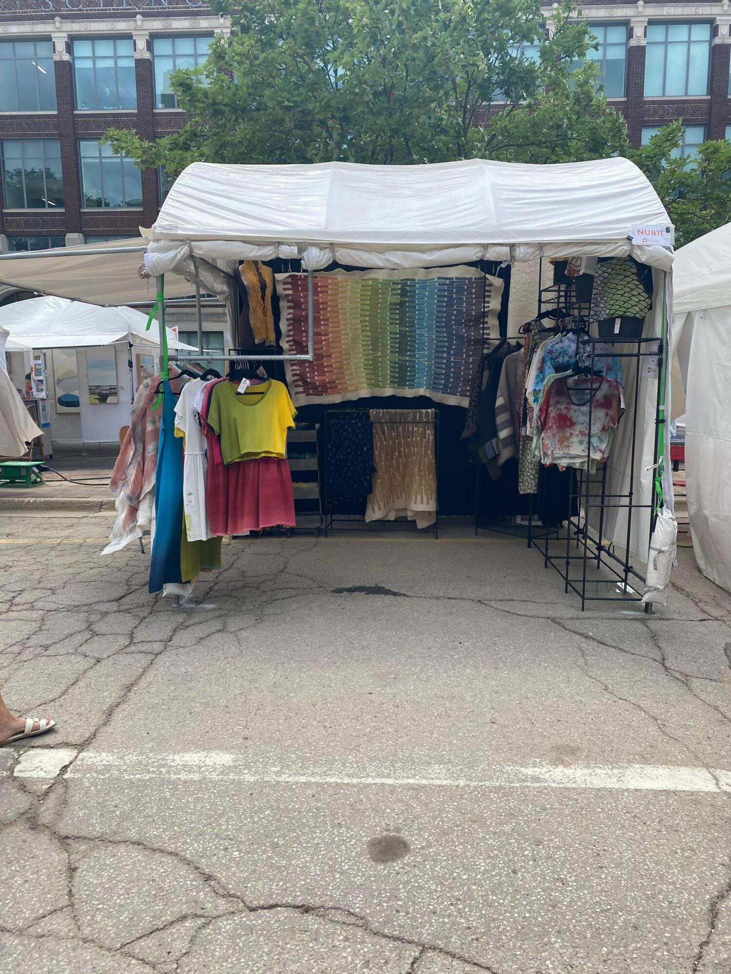 Outdoor market stall with colorful fabric rolls and clothing, shaded by a white canopy, on a paved street.