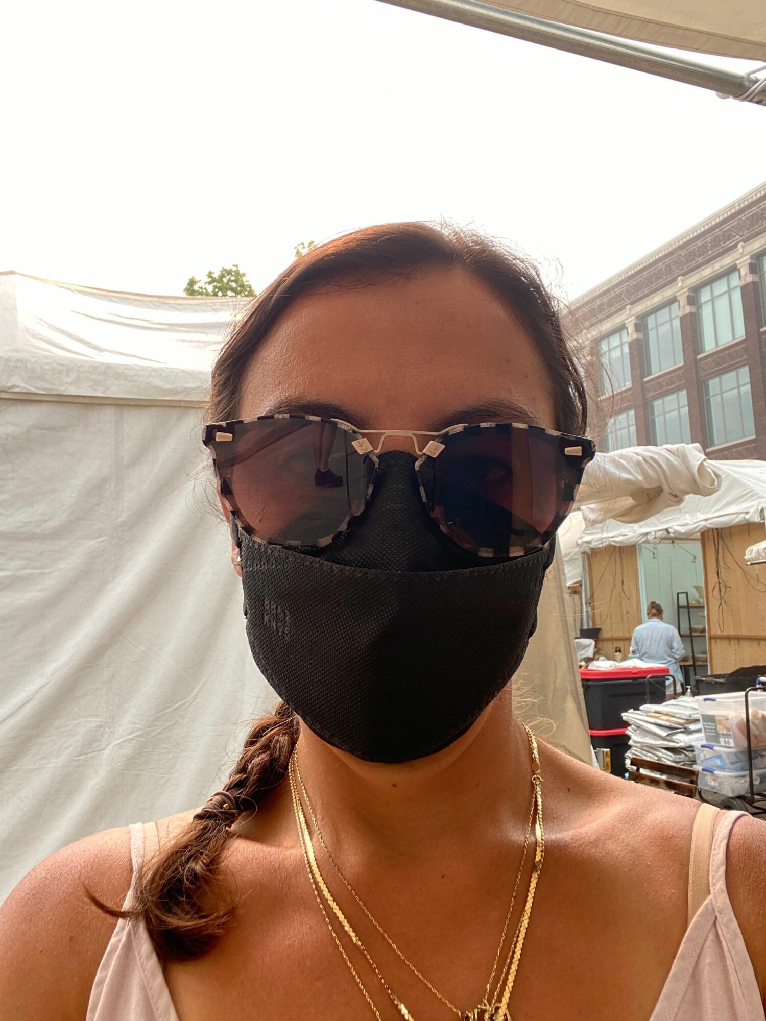 Person wearing sunglasses and a face mask, with a building and outdoor setting in background.
