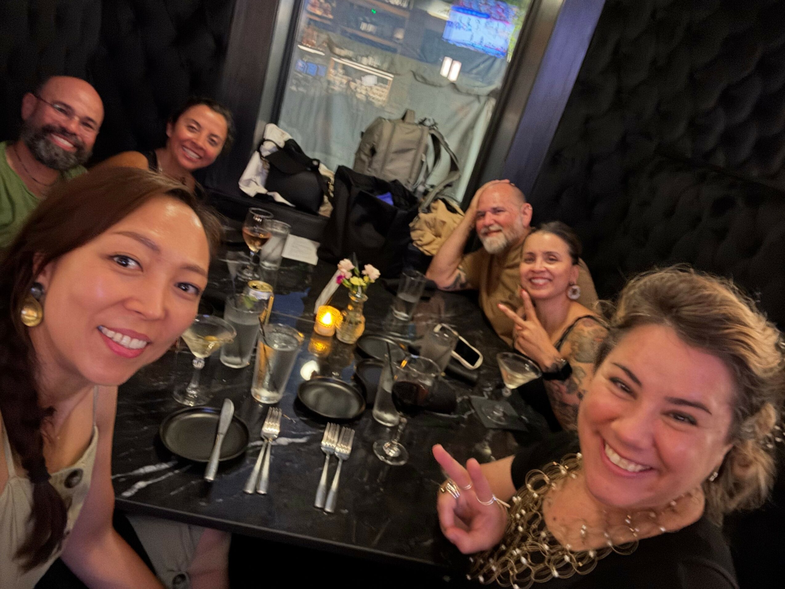 Group of women smiling and taking a selfie at a bar with drinks and glasses on the table.