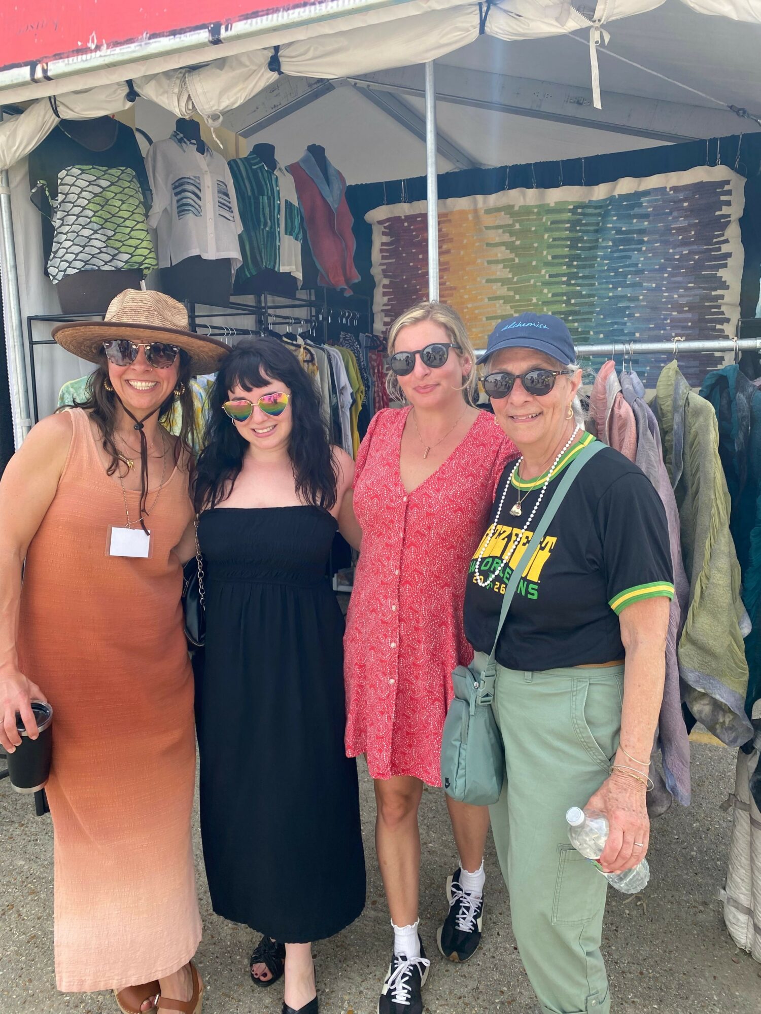 Three people standing together at an outdoor market, smiling, with clothing and textiles displayed behind them.