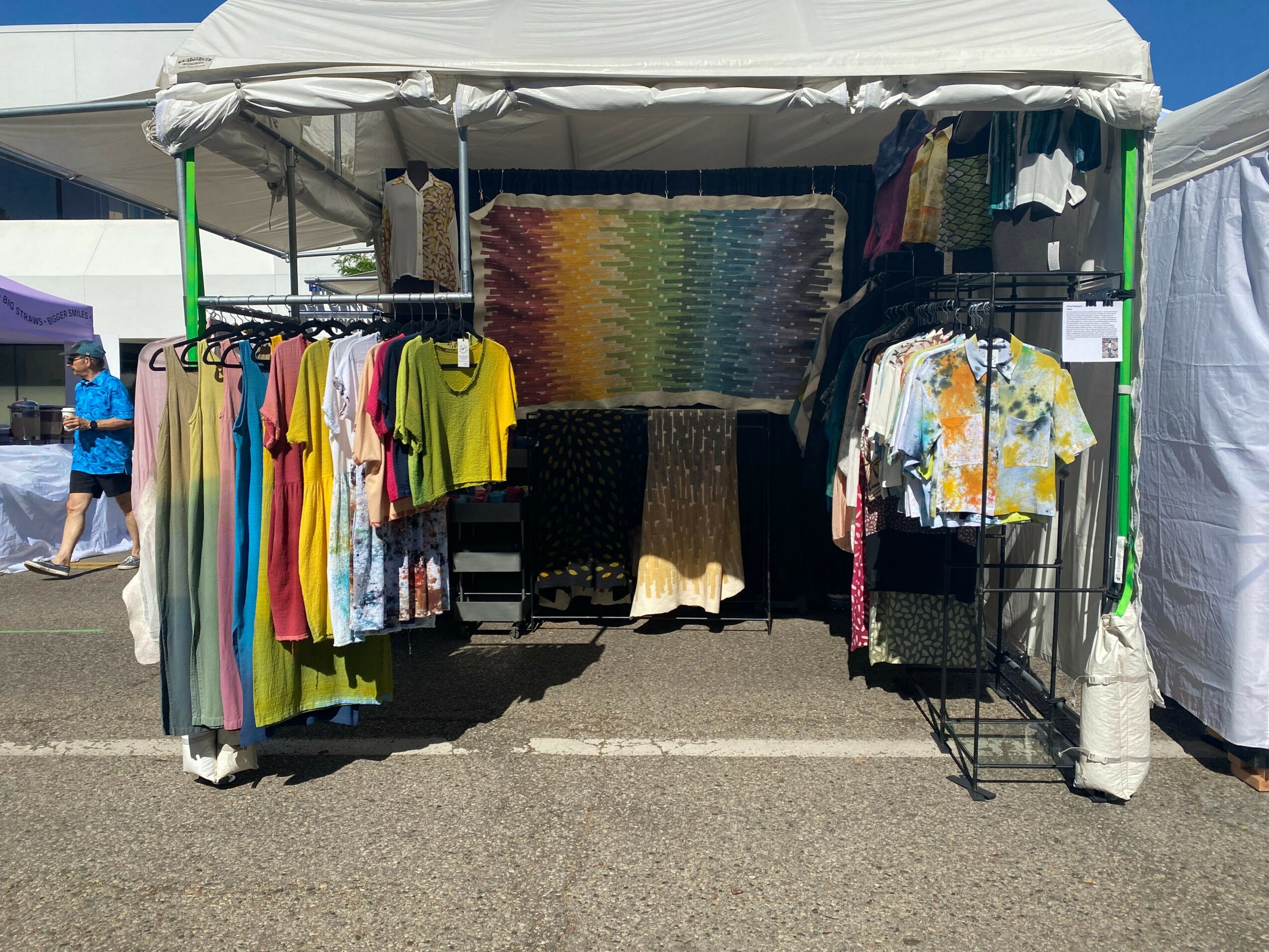 Clothing stall with colorful shirts and dresses displayed on racks under a white canopy tent.