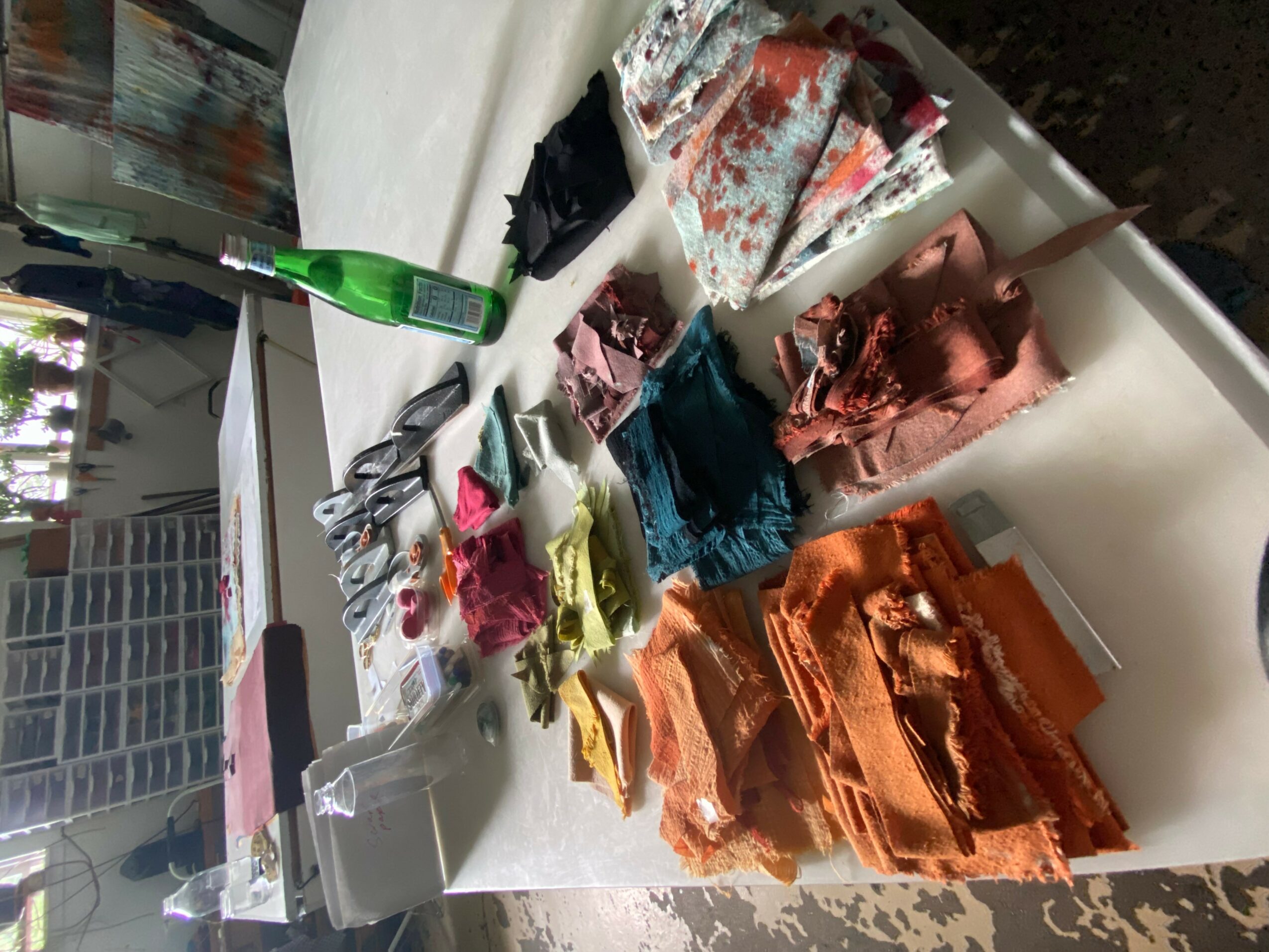 Table with folded clothes, fabric, and a green bottle, viewed from the side, with a window in the background.