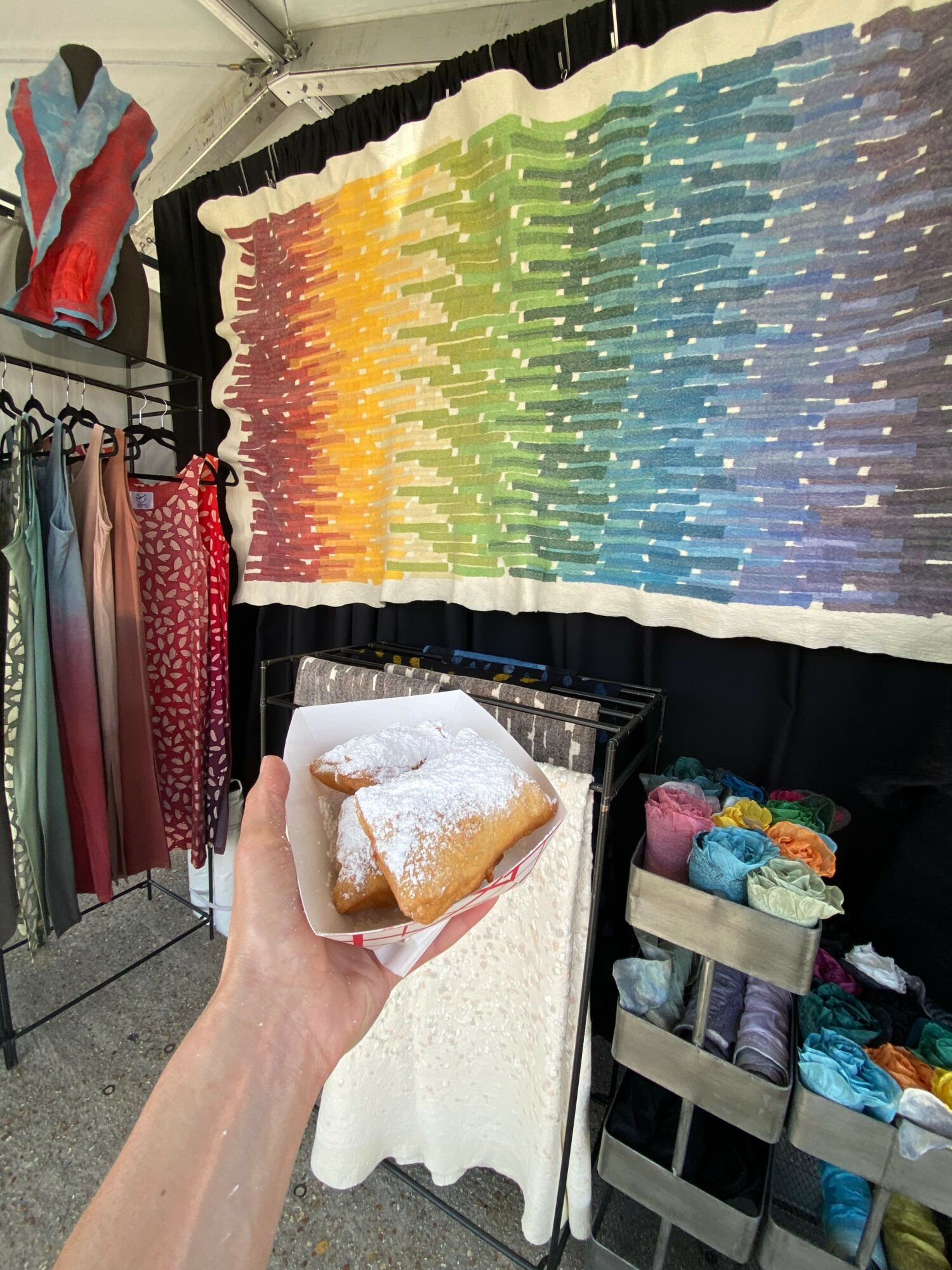Person holding a pastry dusted with powdered sugar, with colorful yarns and a rainbow-colored art piece in the background.