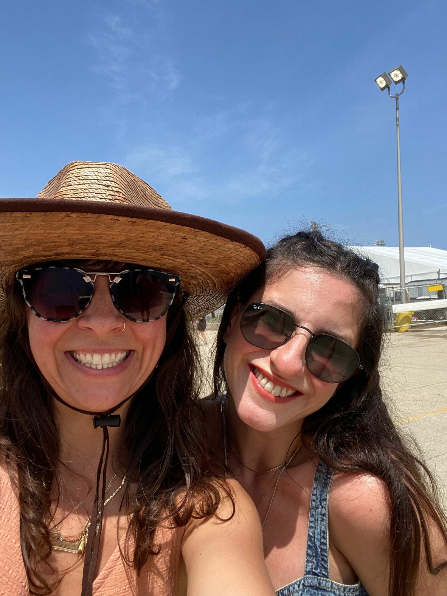 Two women smiling, wearing sunglasses and hats, outdoors on a sunny day.