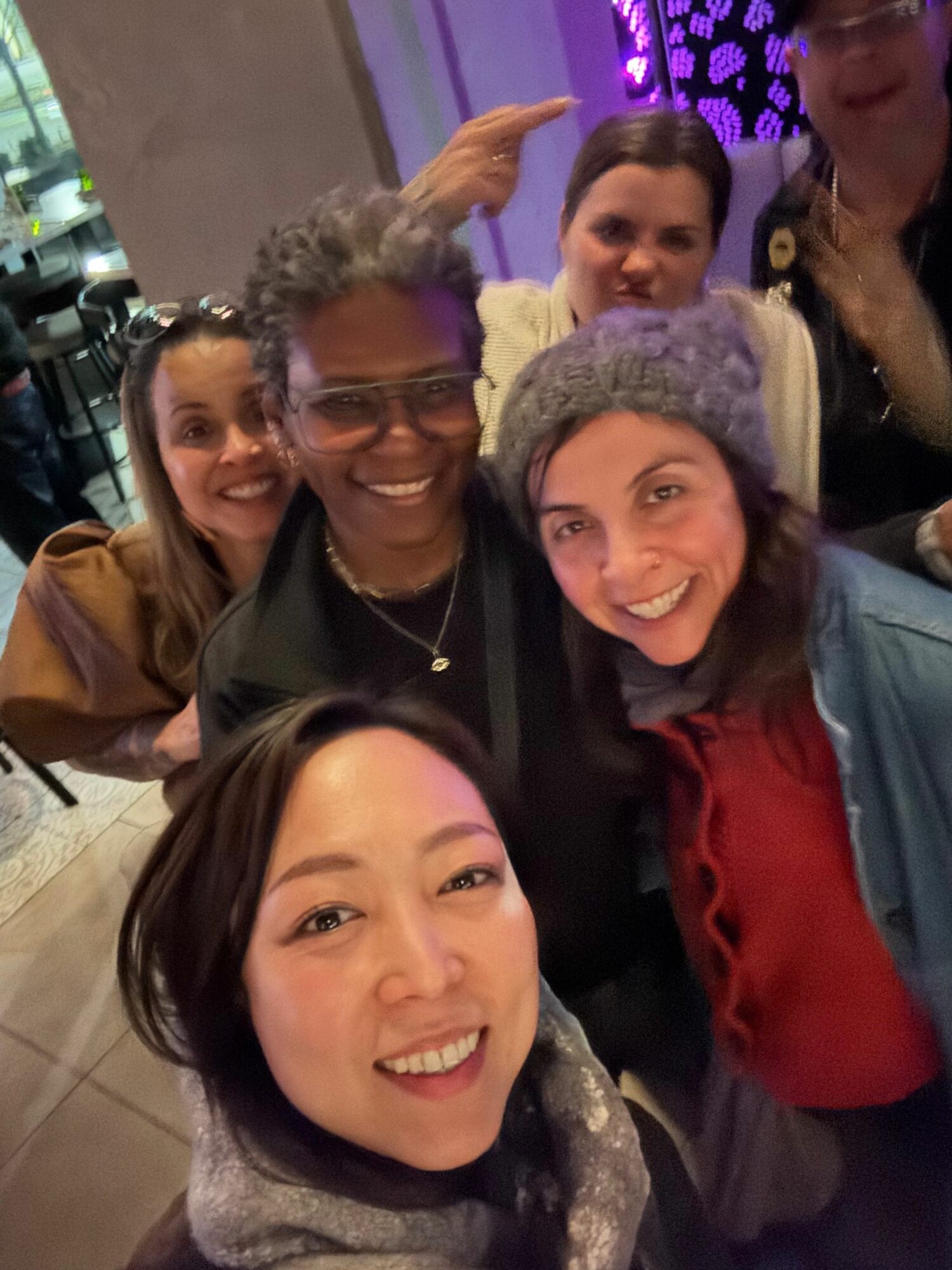 Group of five people smiling and taking a selfie indoors, some wearing glasses and winter clothing.