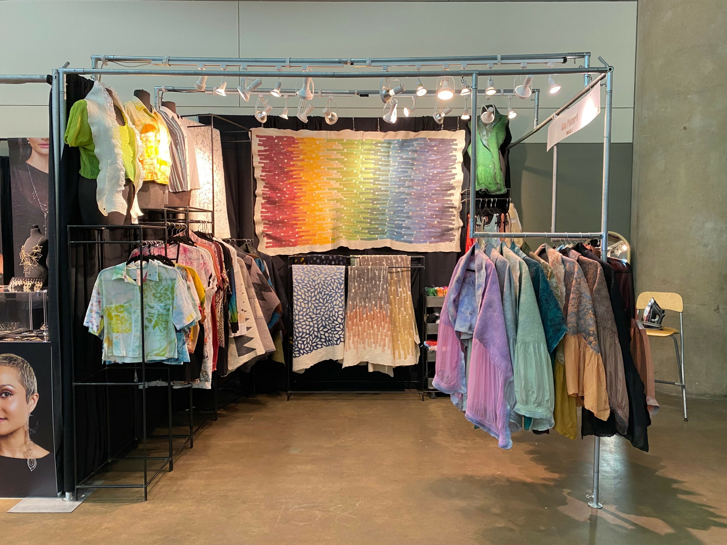 Clothing booth with colorful garments and a rainbow-colored textile hanging above, set in an indoor space.