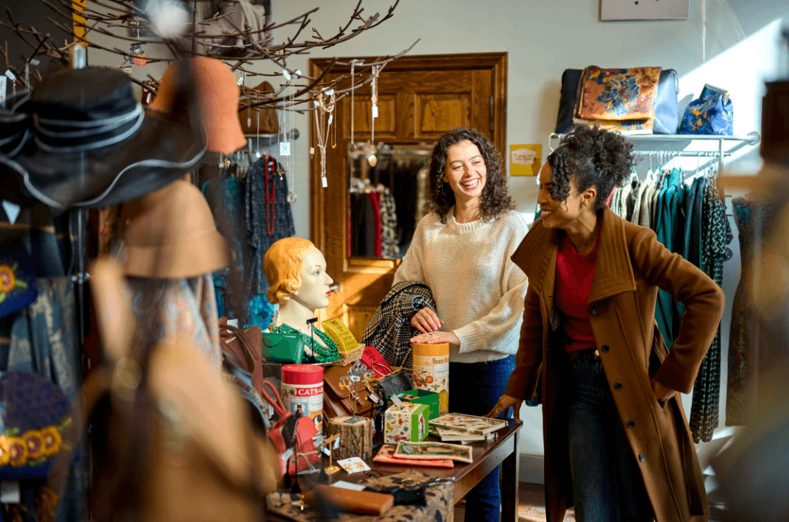 Two women shopping in a clothing store, smiling and talking, with various items on display and a mannequin nearby.