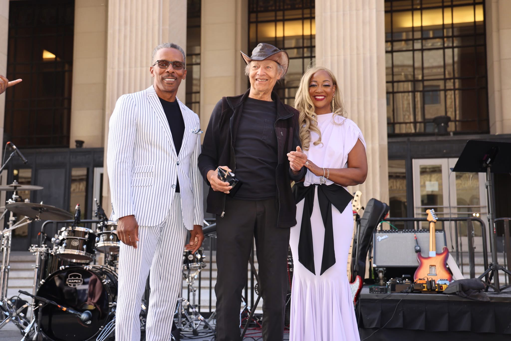Three people standing outdoors in front of a building, smiling, with musical instruments nearby, during daytime.