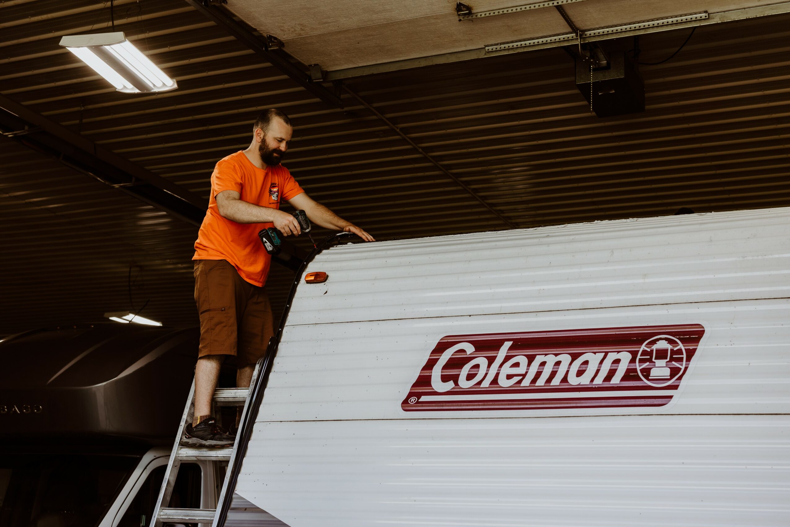 Person standing on a ladder cleaning a Coleman cooler, ceiling with lights visible above, indoor setting.