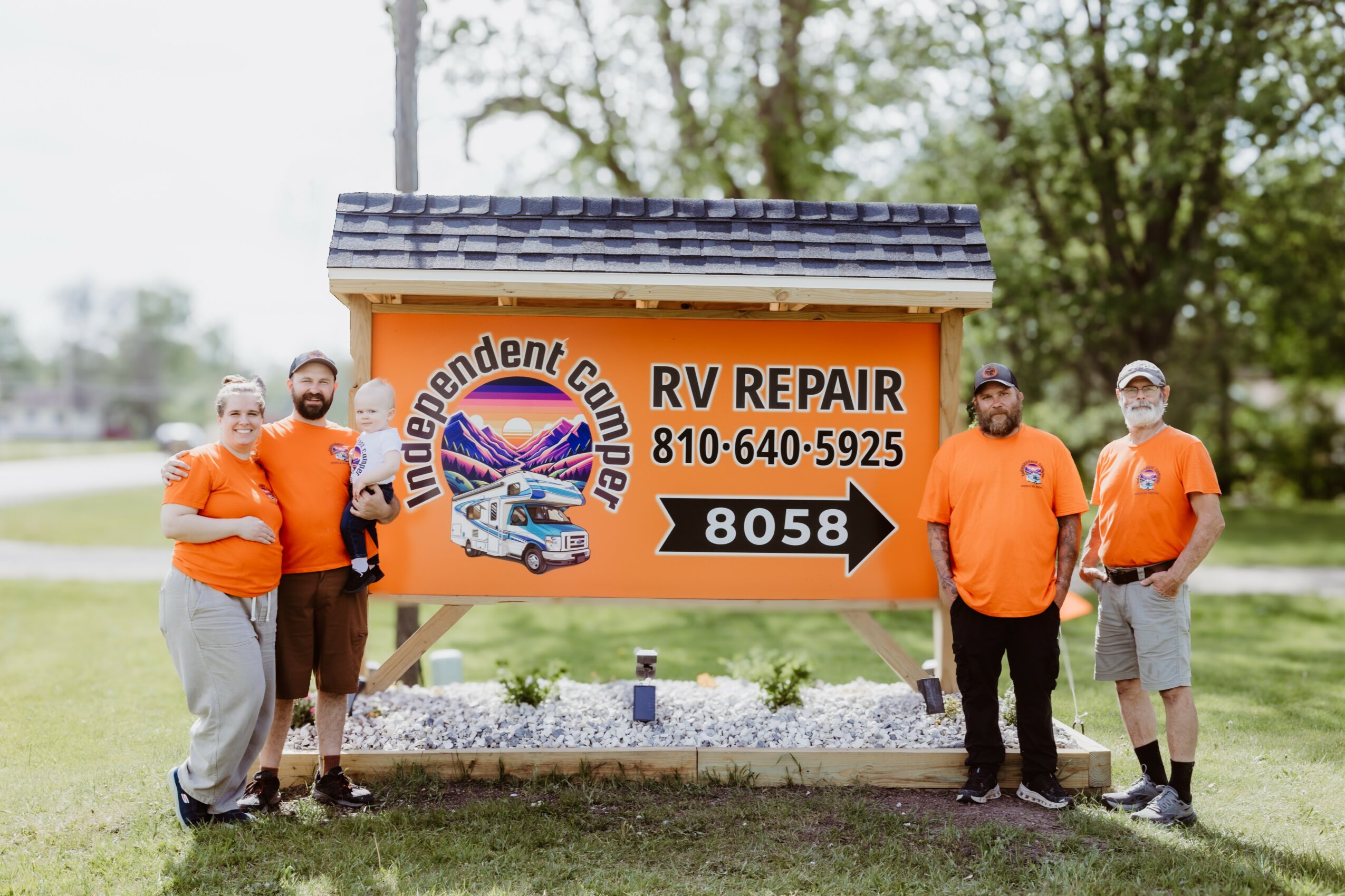 Five people in orange shirts stand beside a sign for RV repair, outdoors with trees in the background.