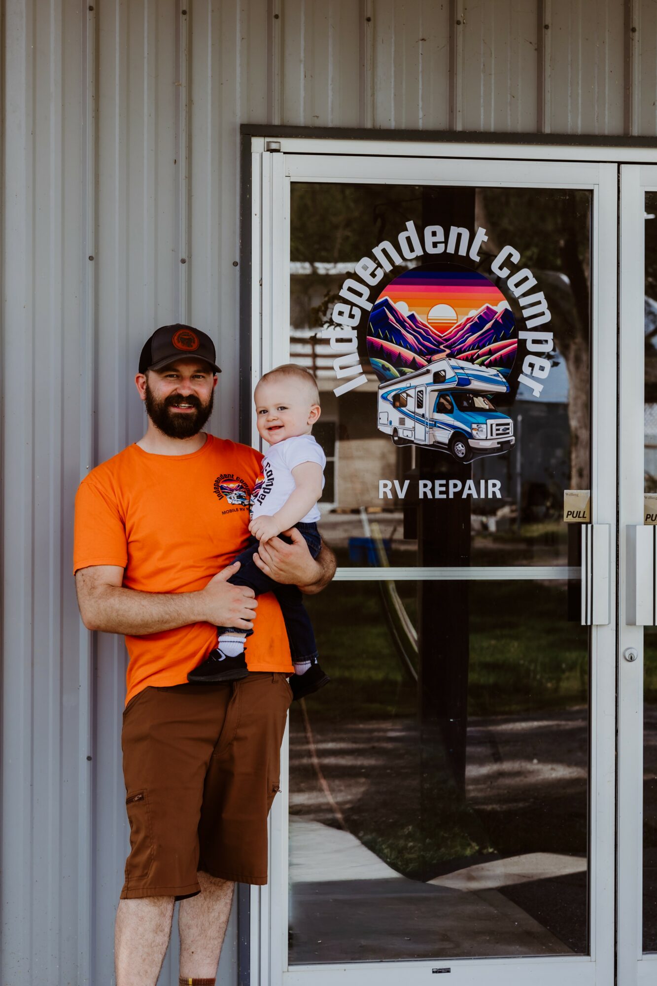 Man holding a baby in front of a glass door with a logo for an independent company and RV repair.