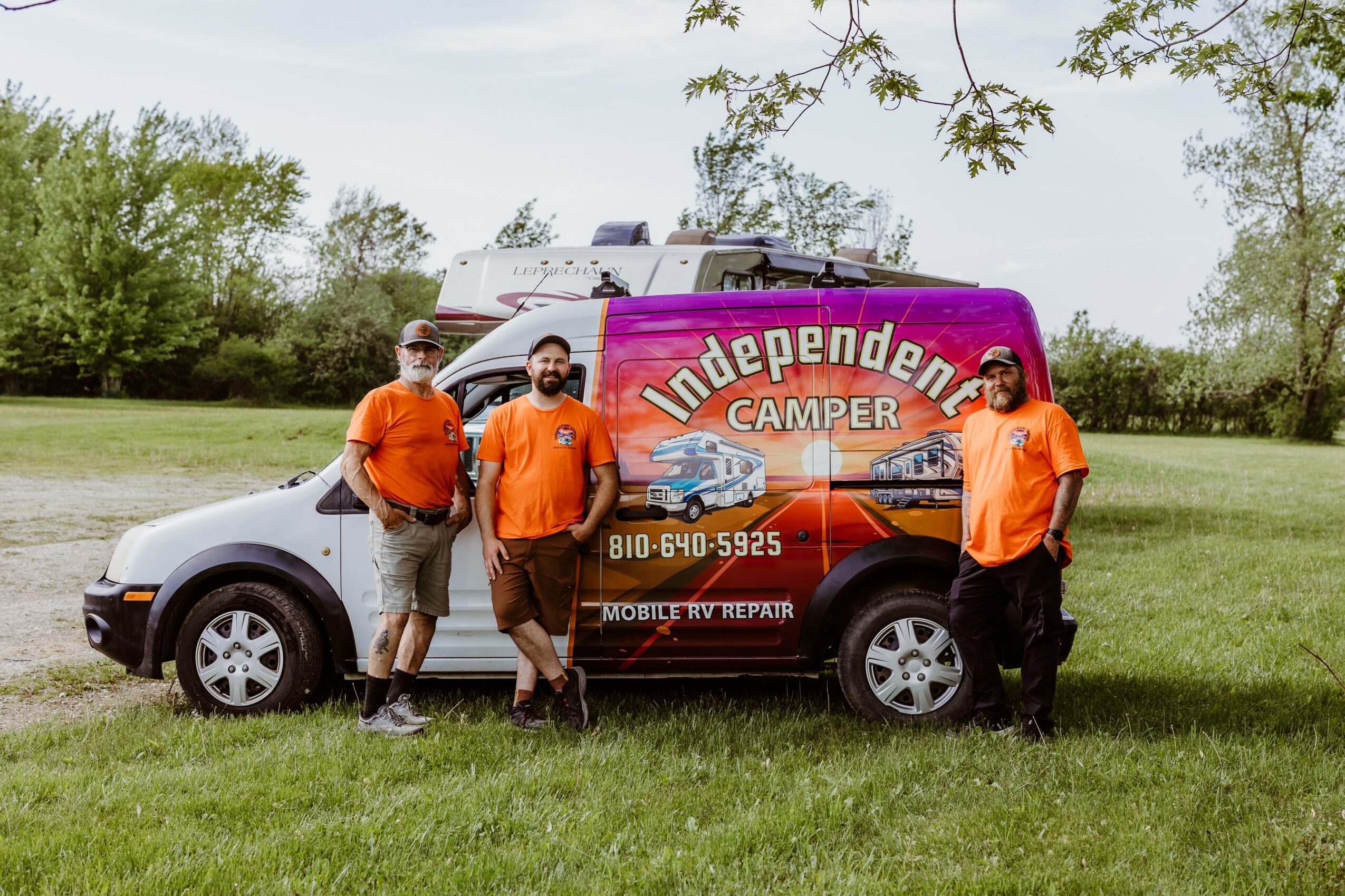 Four people stand beside a camper van with a purple and orange design, in a grassy outdoor area with trees.
