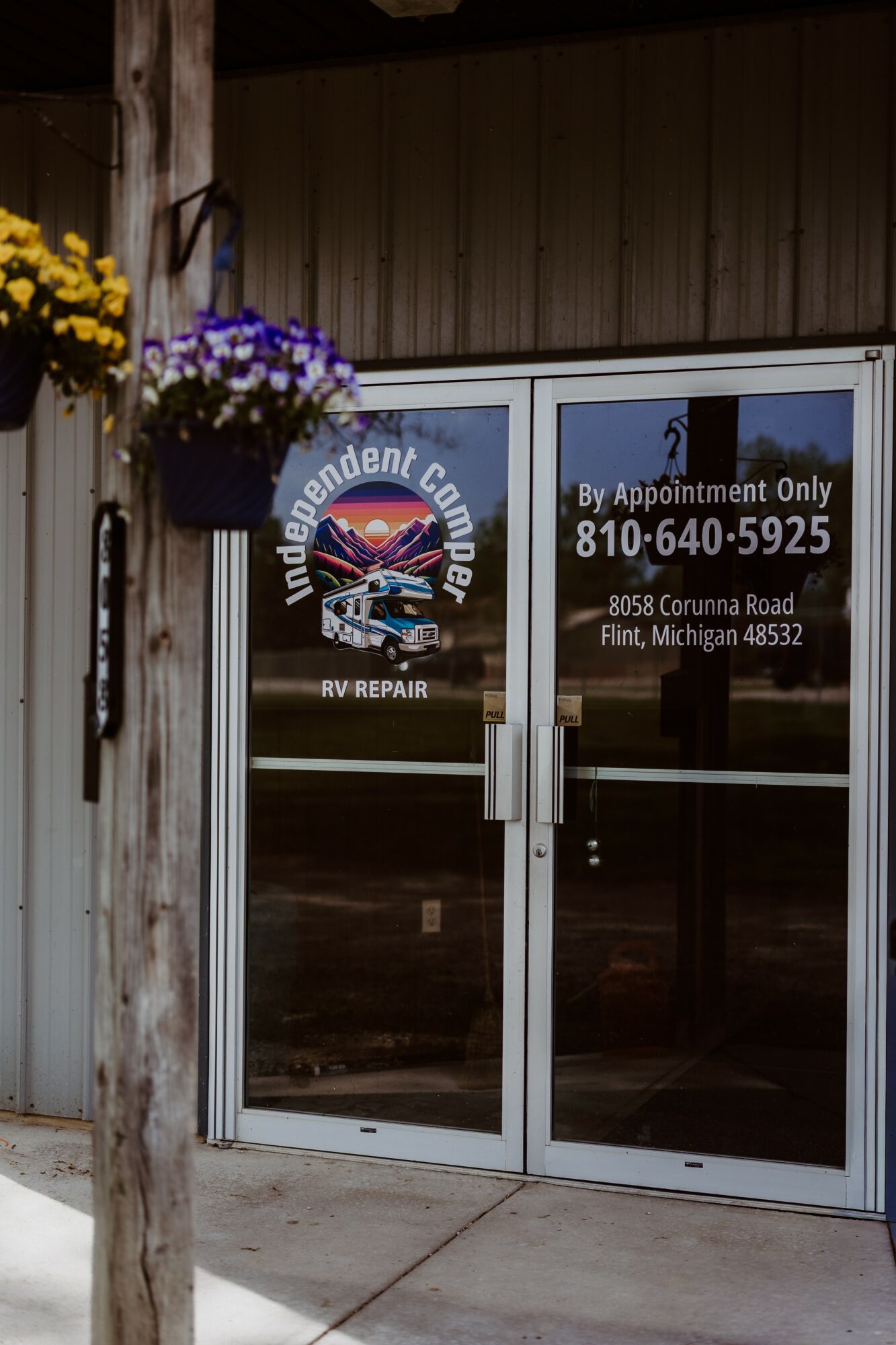 Glass door with signage for an independent auto repair shop, flowers hanging on the left, and a wooden wall above.