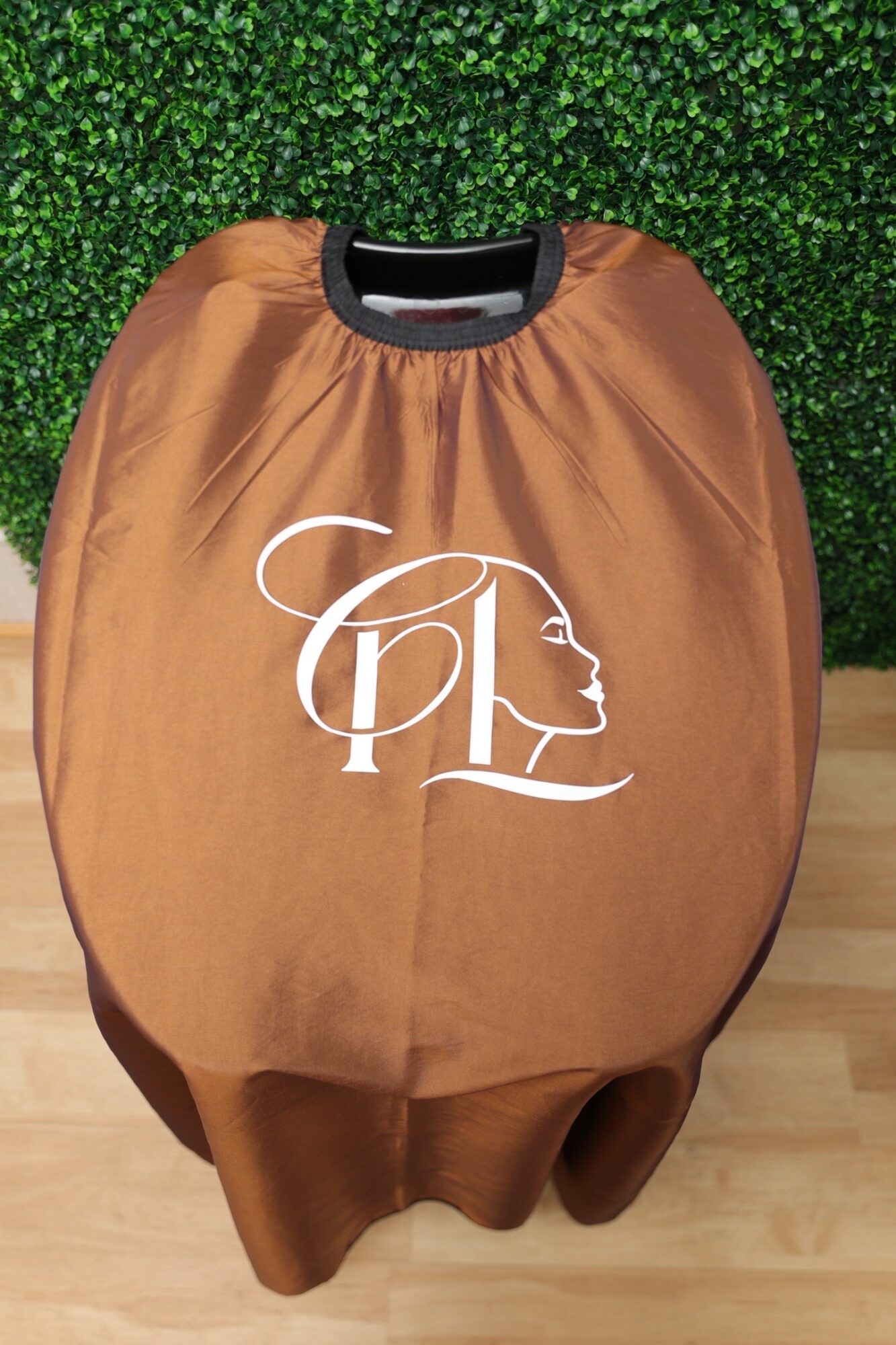 Brown salon cape with a white logo of a woman's face and hair design, hanging against a green leafy background.