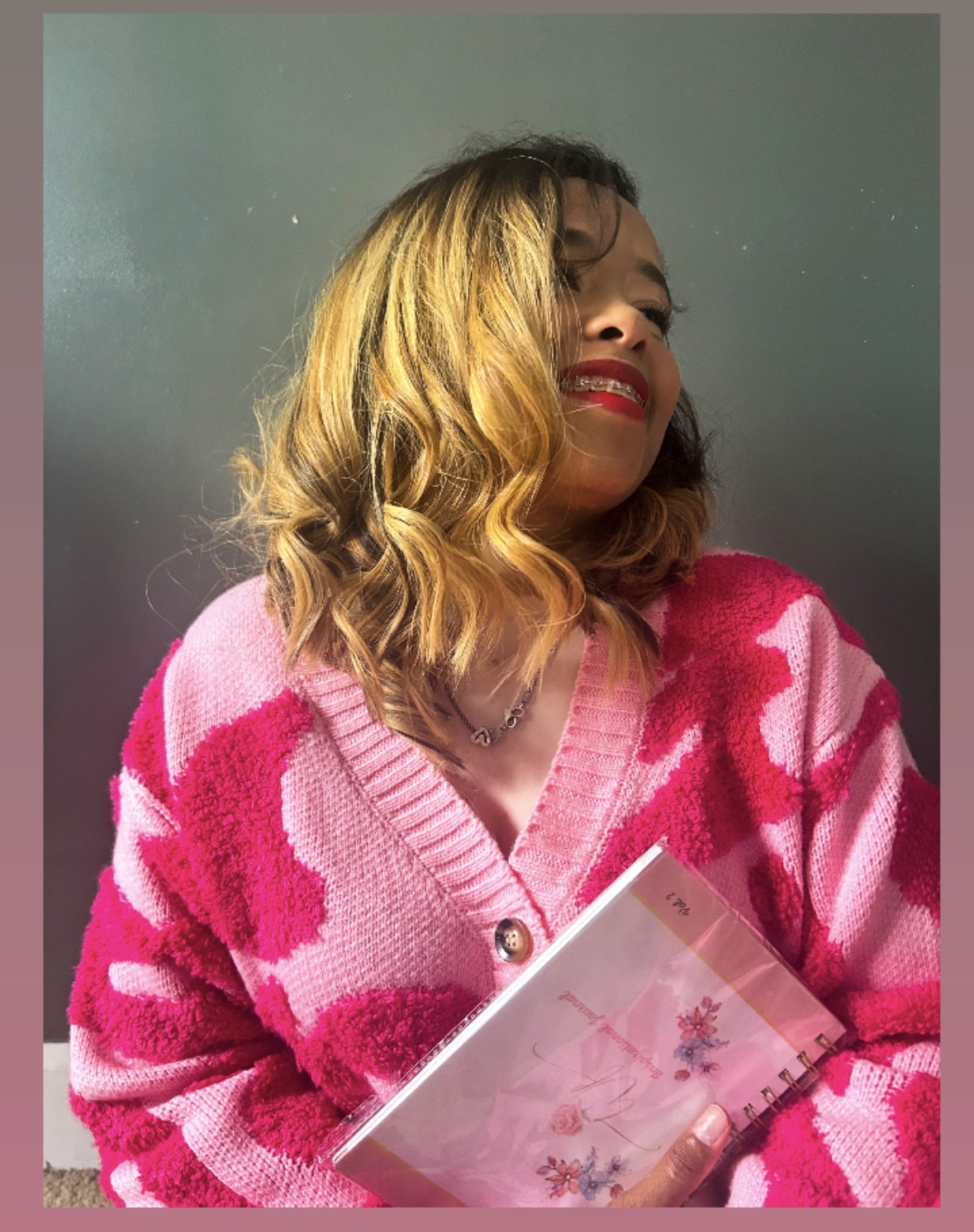Woman with wavy blonde hair wearing a pink and red patterned sweater, holding a notebook, smiling with eyes closed.