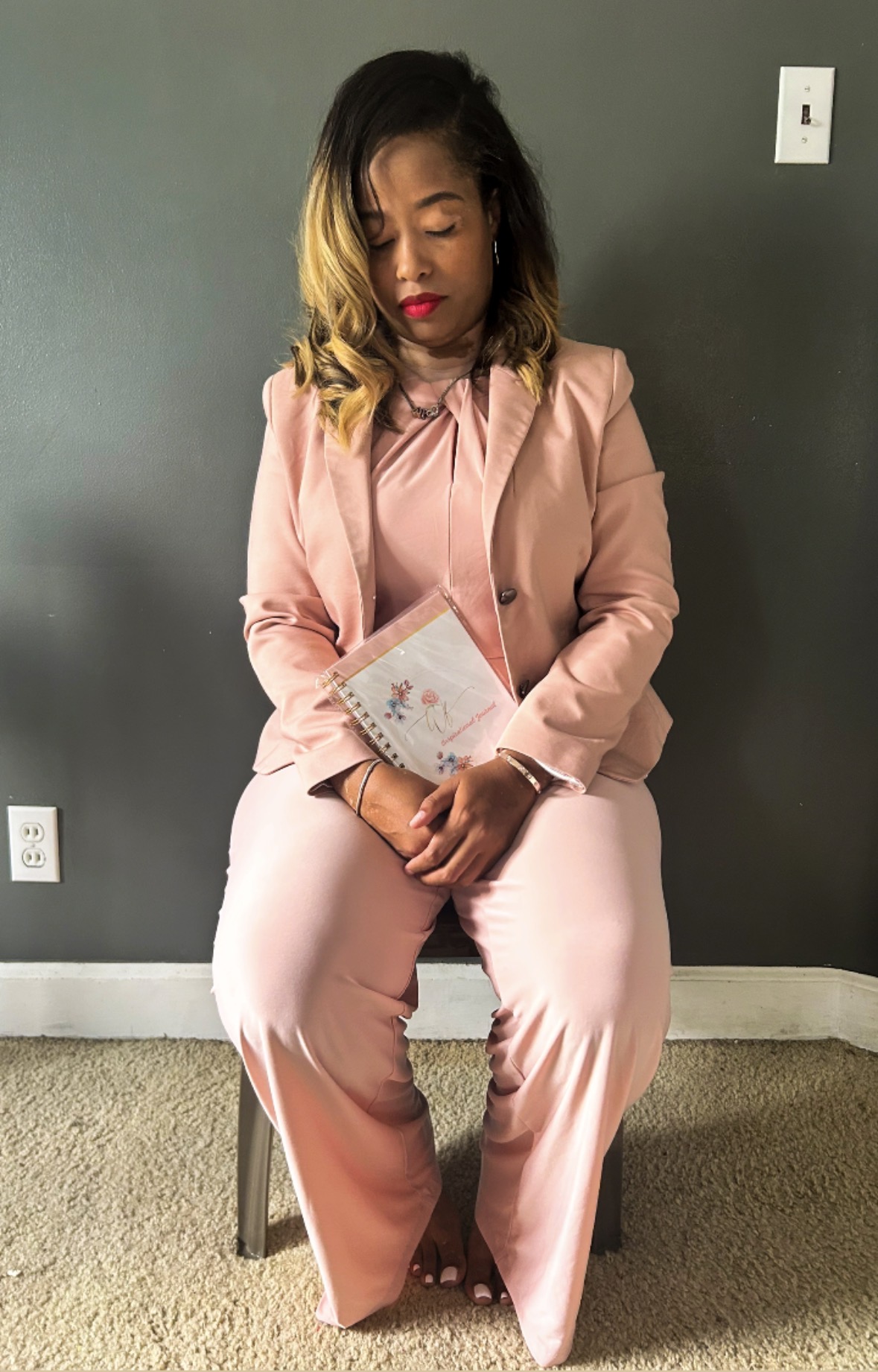 Woman in pink suit sitting on a chair holding a book, with a gray wall background.