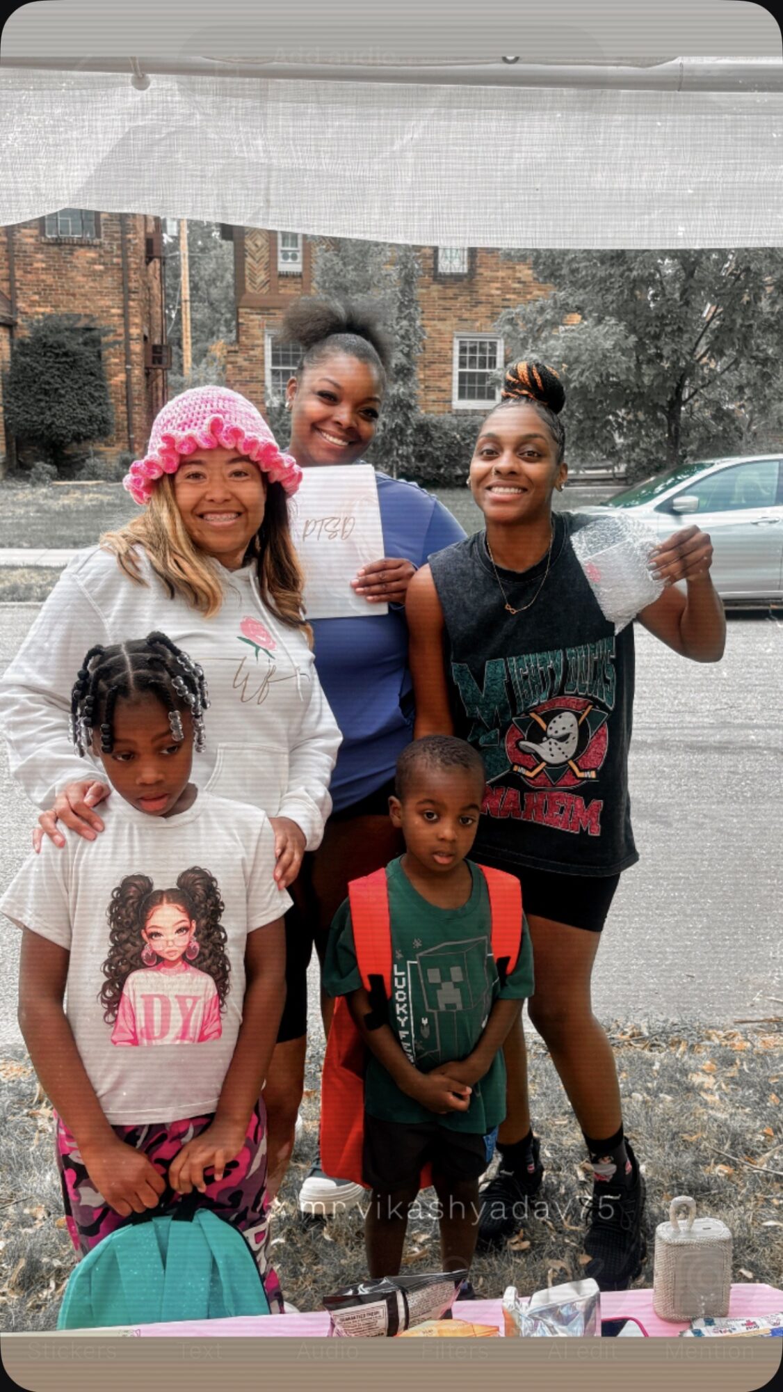 Four children and a woman standing outdoors on a street, smiling, with trees and buildings in the background.