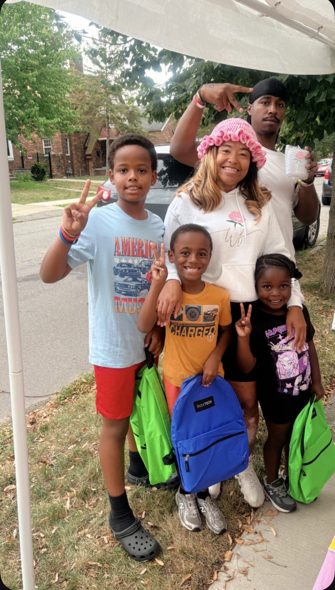 Group of five children and one adult outdoors, smiling, with backpacks, under a white canopy, trees and a street in background.