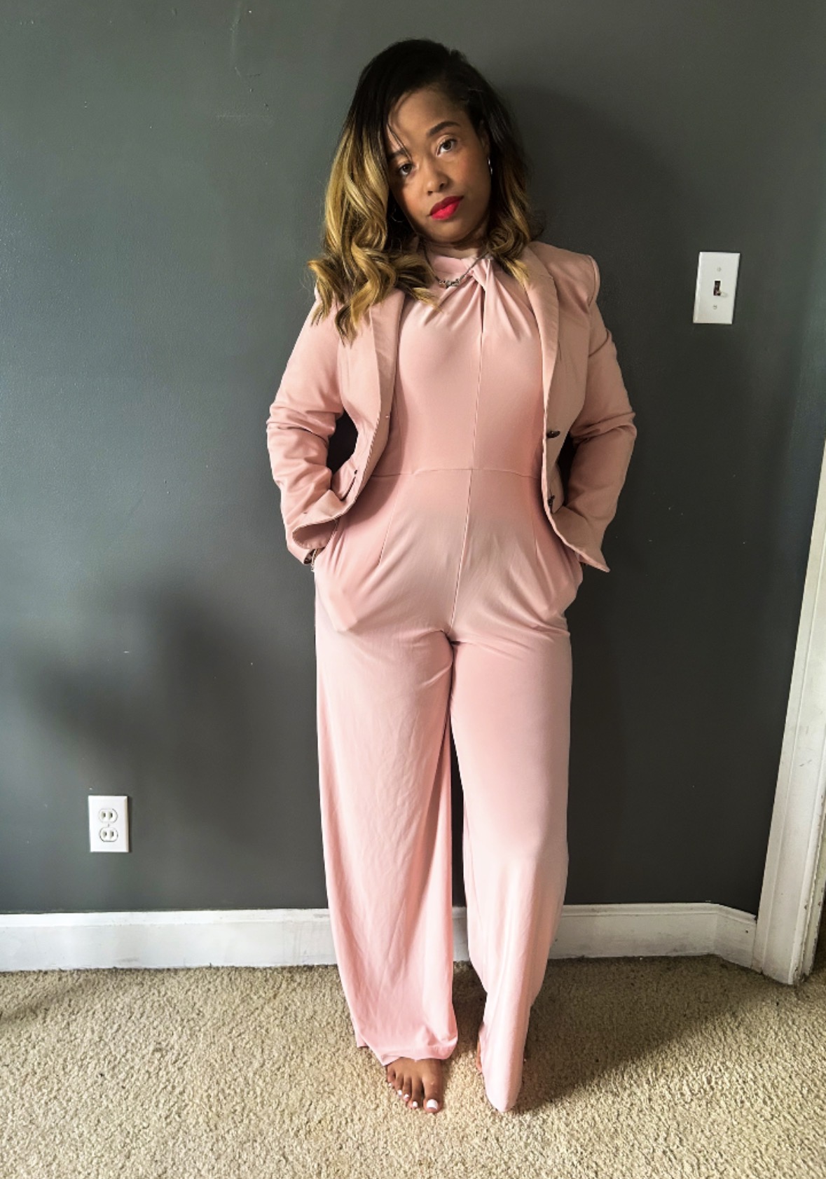 Woman in pink jumpsuit standing indoors with hands in pockets, dark gray wall background, beige carpet floor.