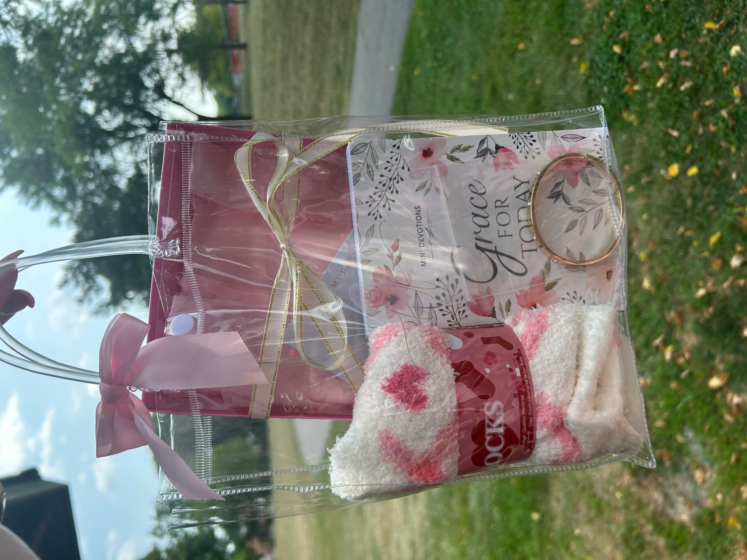 Clear gift bag with pink ribbon, containing a small towel with a pink heart, a pink candle, and a card, outdoors.