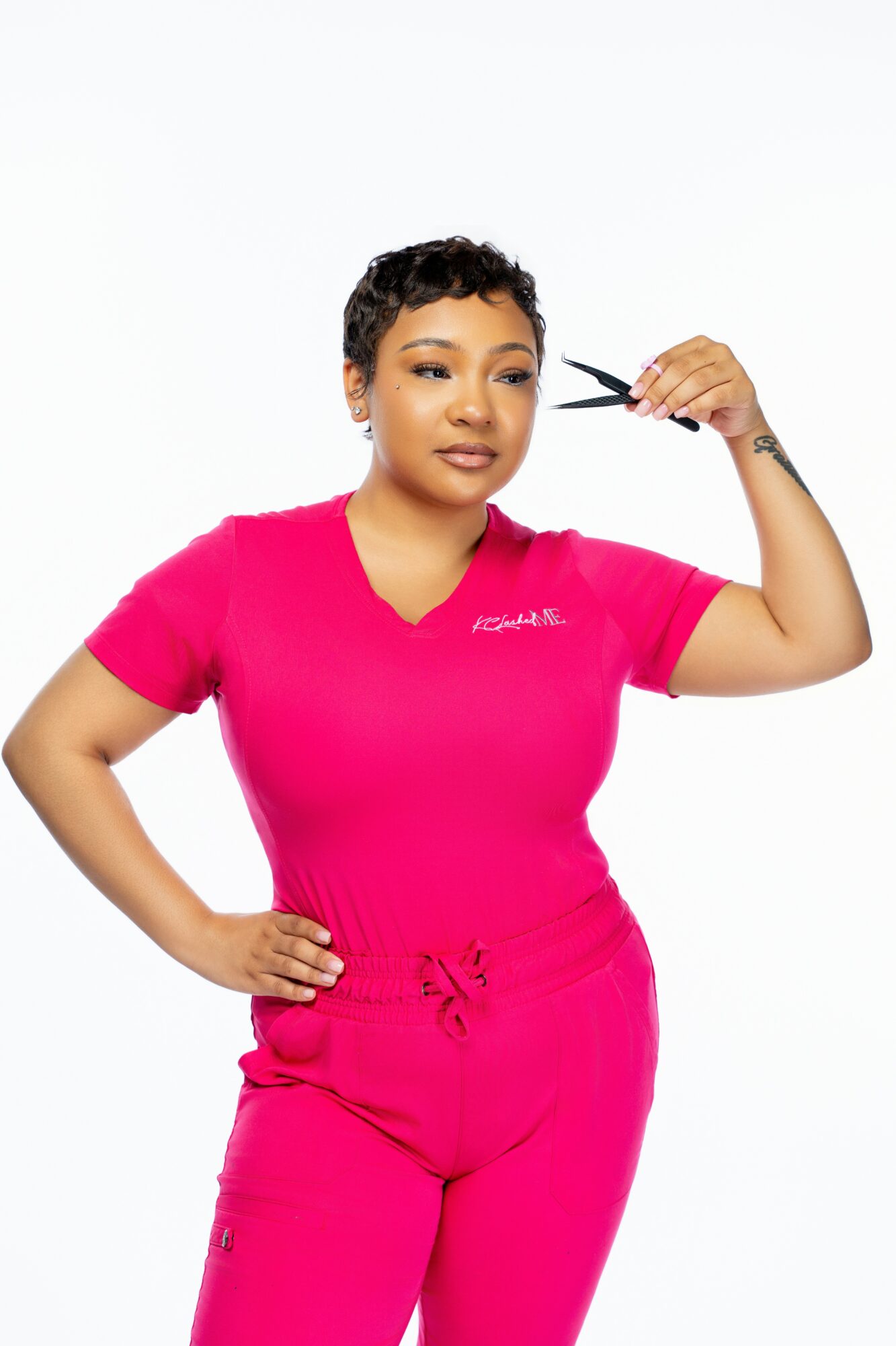 Woman with short curly hair in pink scrubs holding scissors near her head, smiling, against a white background.